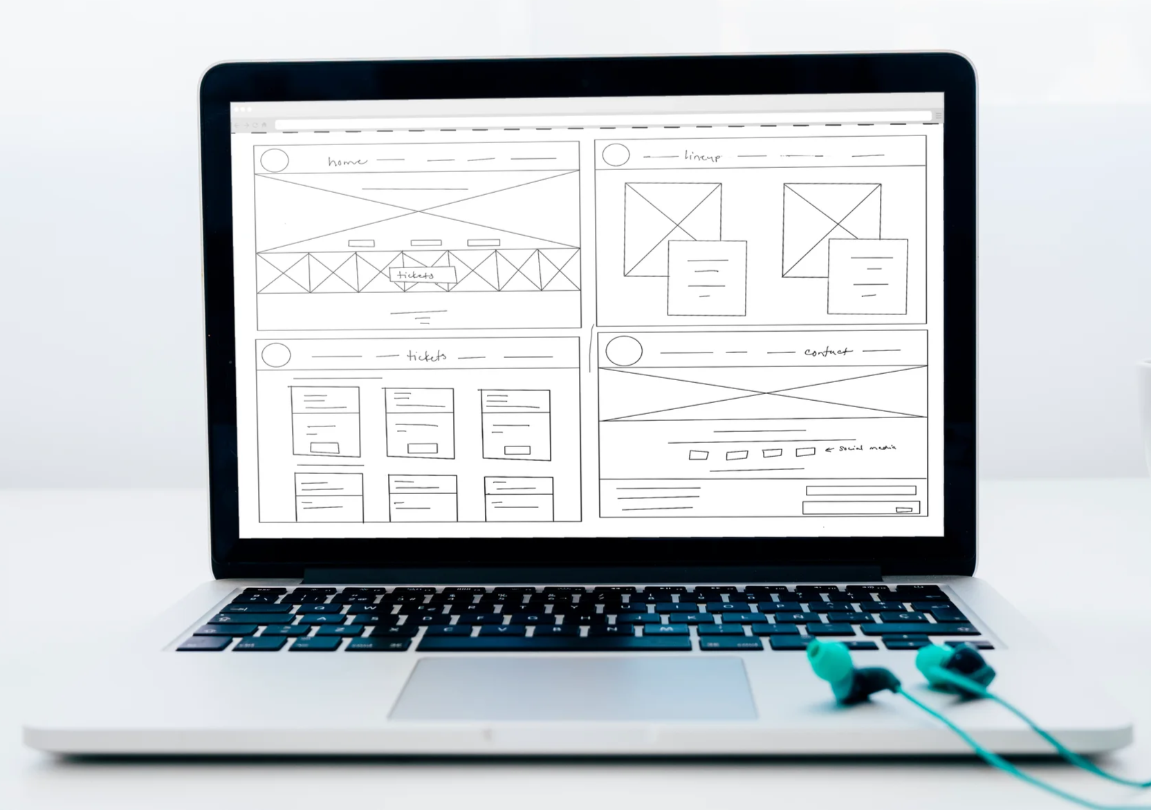 Laptop on a white surface displaying a rough sketch of a website wireframe with different sections labeled 'home', 'linear', 'tickets', and 'contact' and an illustration of earbuds next to it.