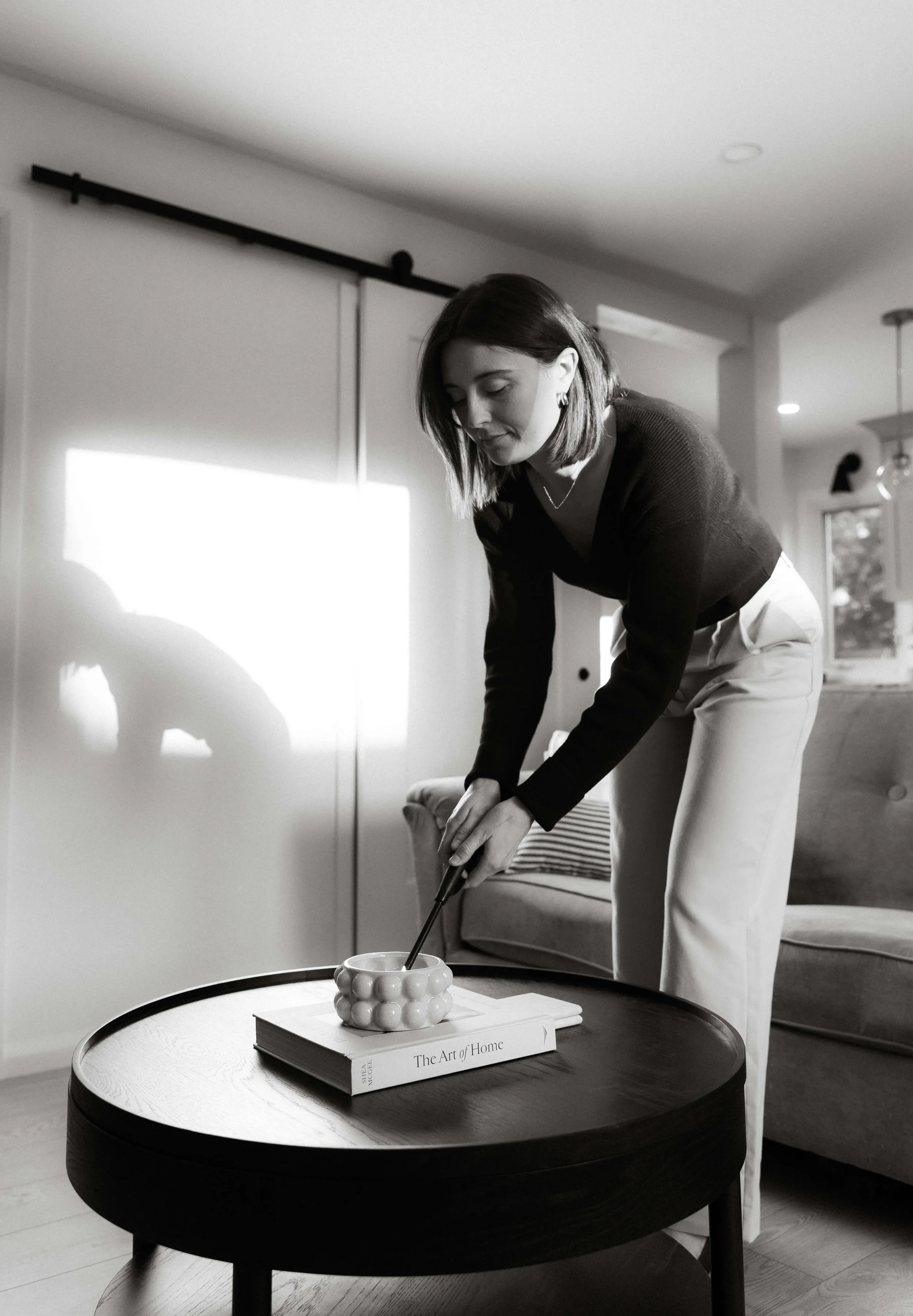 A woman with shoulder-length dark hair, wearing a dark long-sleeve top and light-colored pants, is bending over and stirring a cup on a small round table. The table has a book titled 'The Art of Home' and a decorative ceramic bowl. The setting is a cozy living space with a couch, window, and a sliding door with a black track in the background.