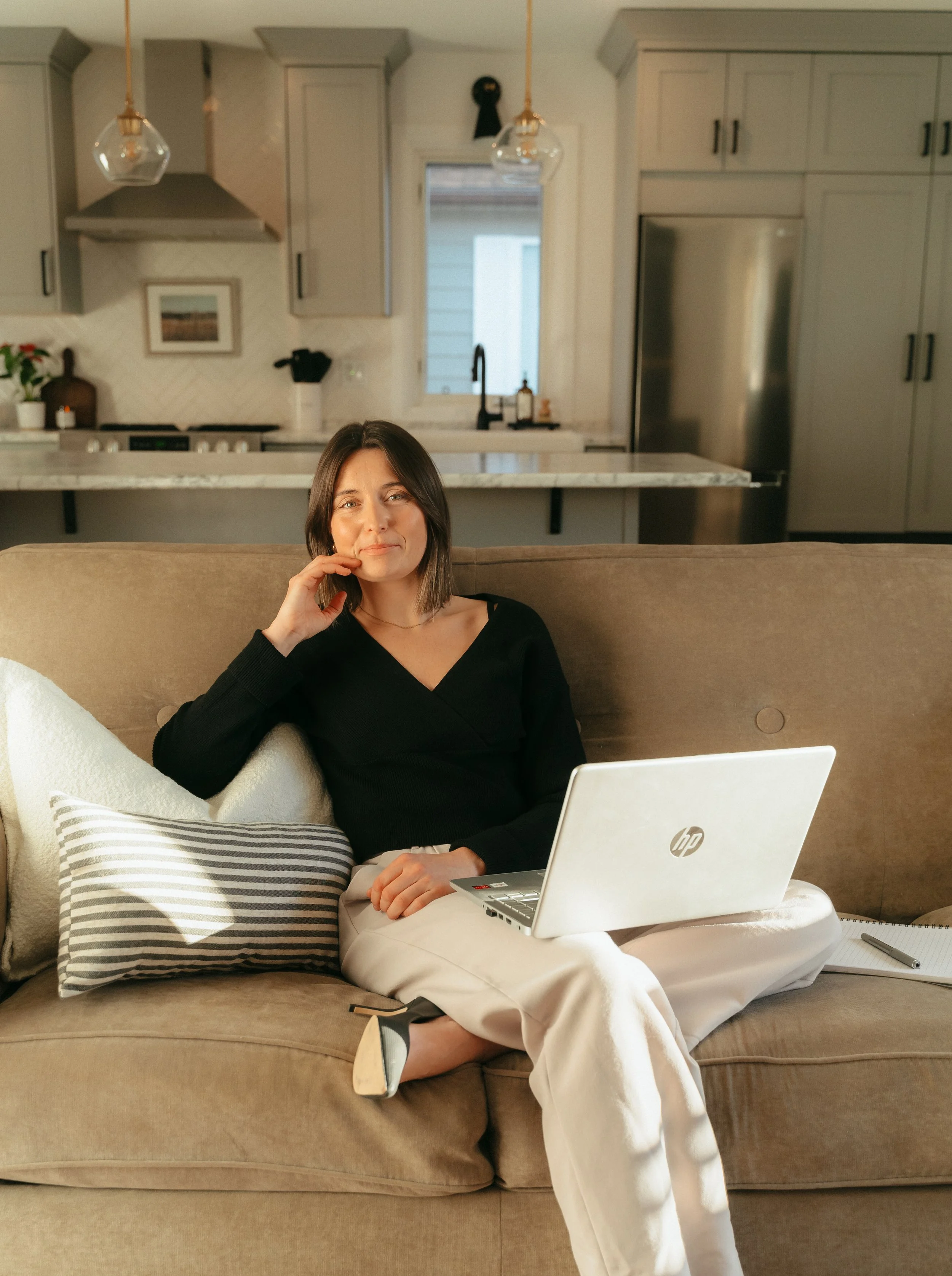 A woman sitting on a beige sofa in a kitchen, working on a laptop and looking at the camera in a well-lit modern home interior.