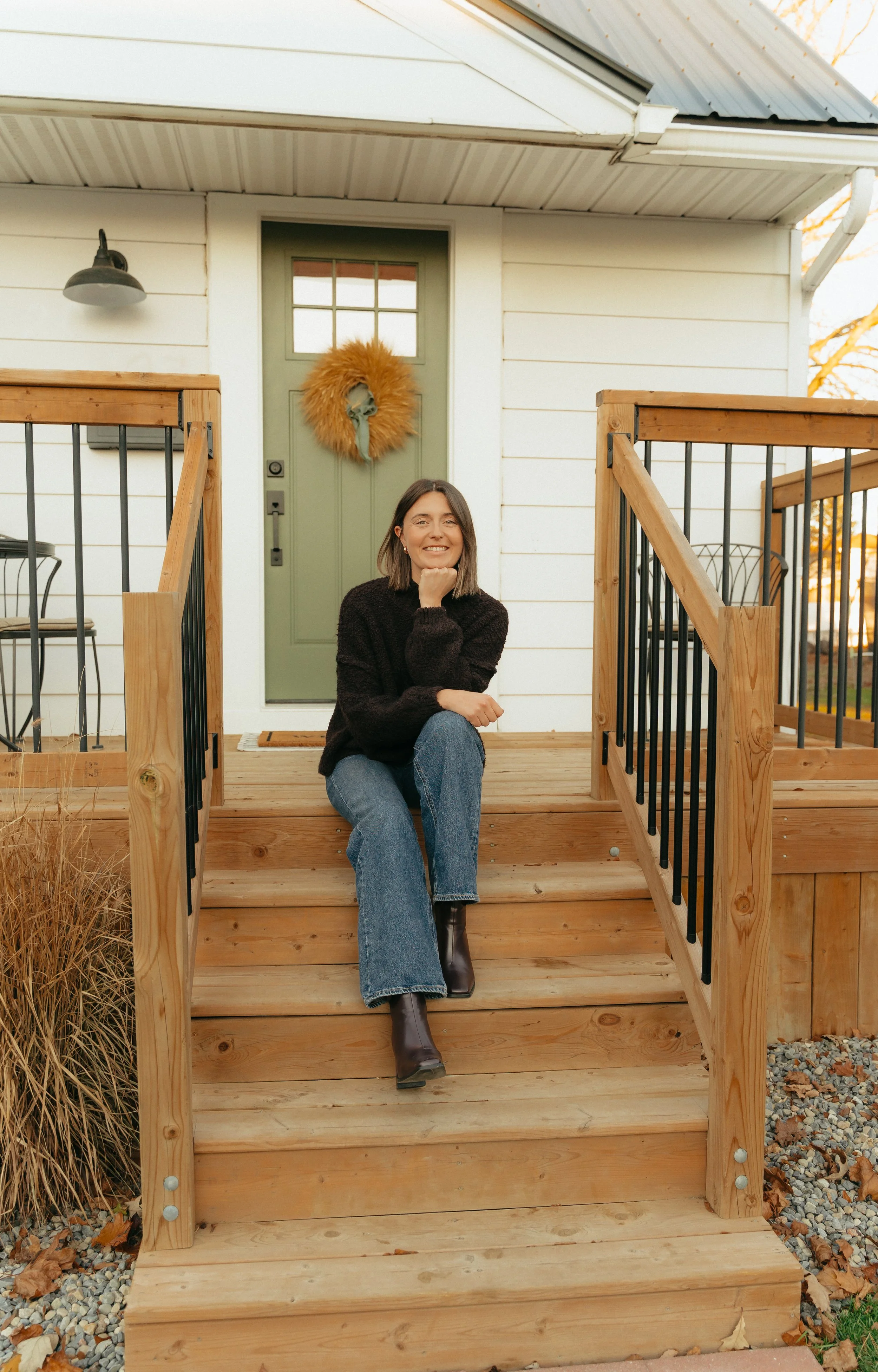 Smiling woman sitting on wooden porch steps of a house with white siding, green door with a yellow wreath, and black metal railing, during fall season.