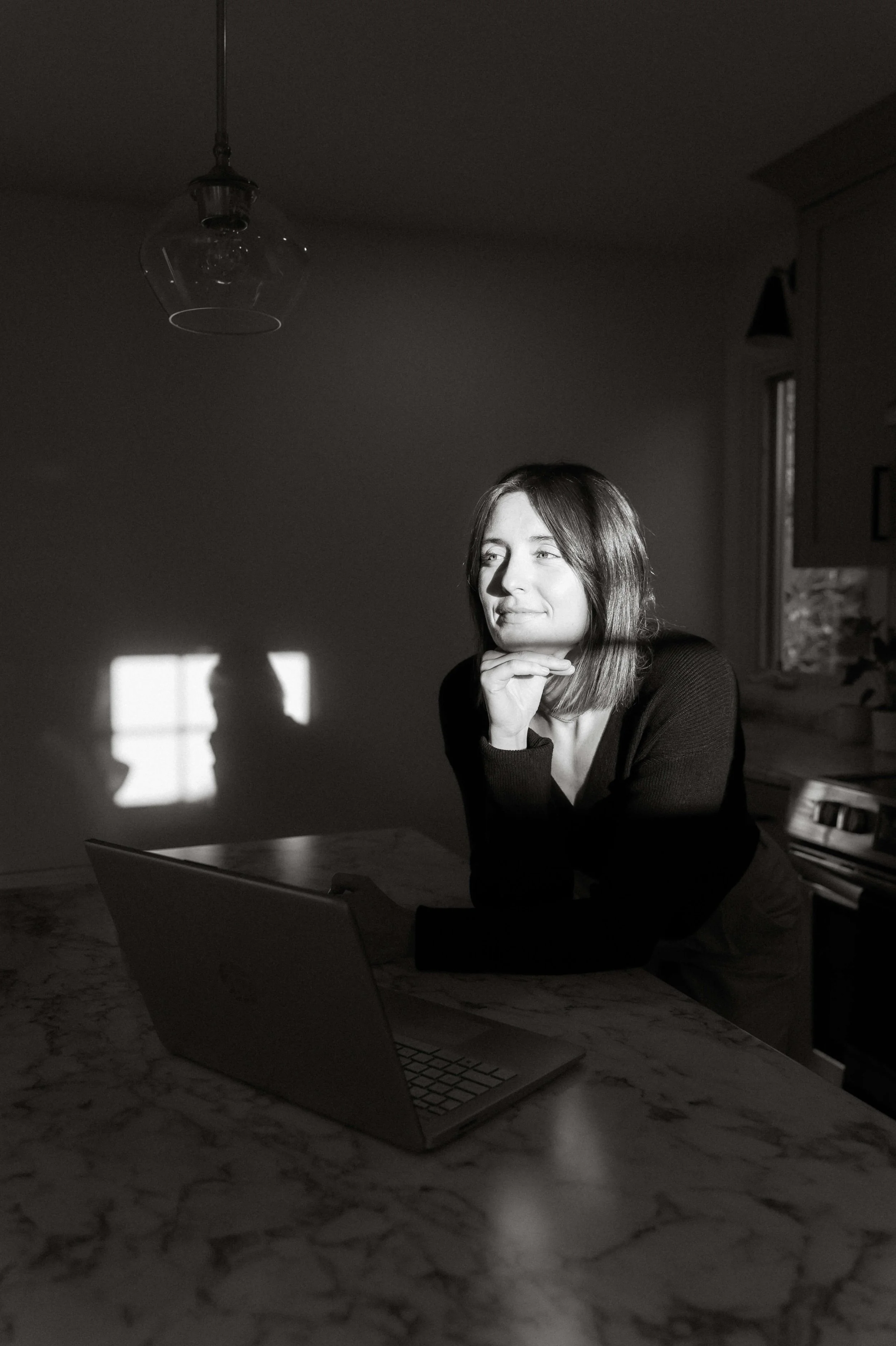 Black and white photo of a woman with shoulder-length hair sitting at a table with her chin resting on her hand. She is smiling slightly and appears to be looking at something outside the frame. The scene is lit by natural light coming through a window, casting shadows on the wall behind her, including her shadow on the wall.