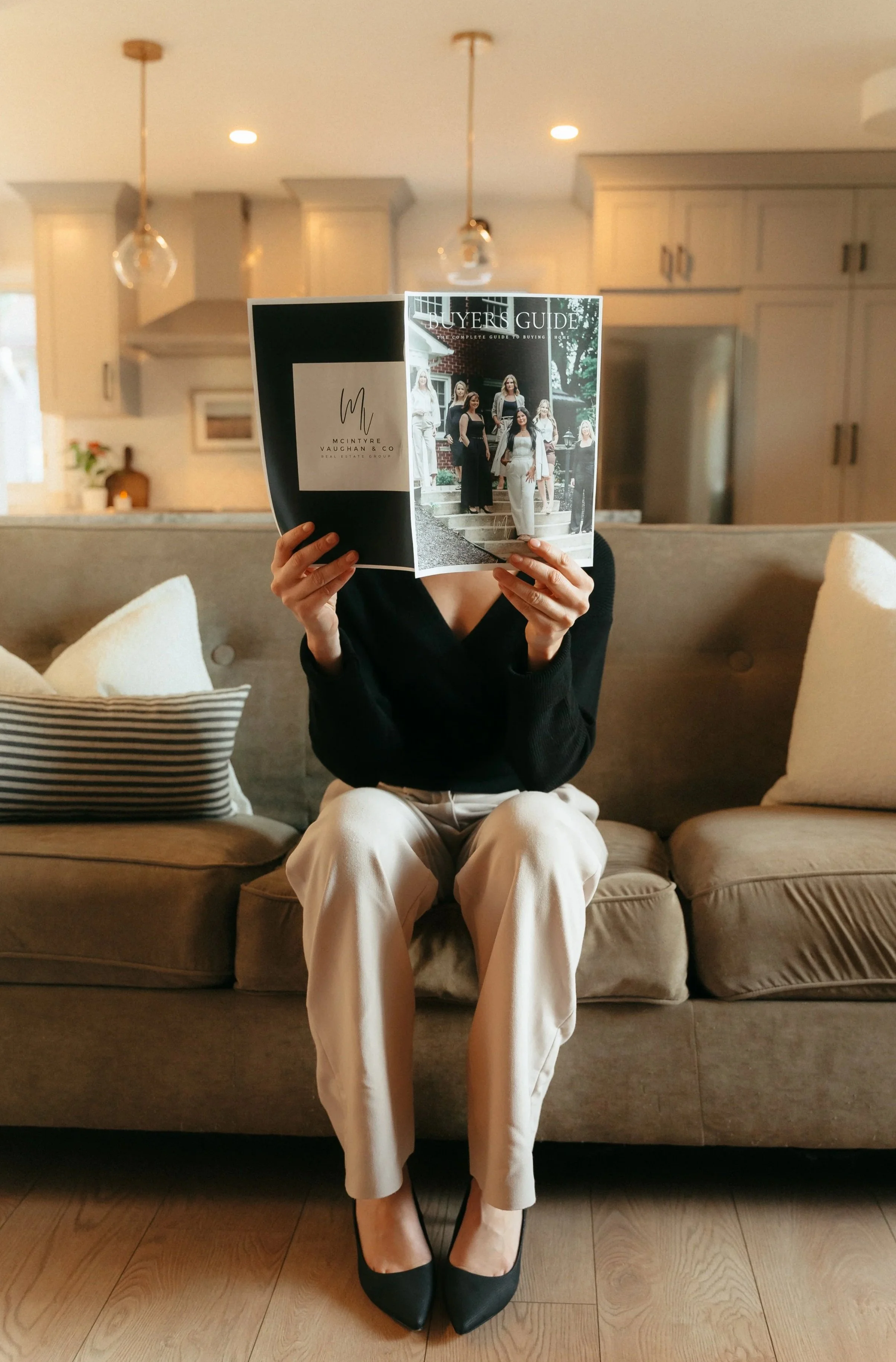 Person sitting on a beige sofa with striped and solid pillows, reading an open magazine that covers their face, in a cozy living room with wooden floors, beige and white furniture, and a kitchen in the background.