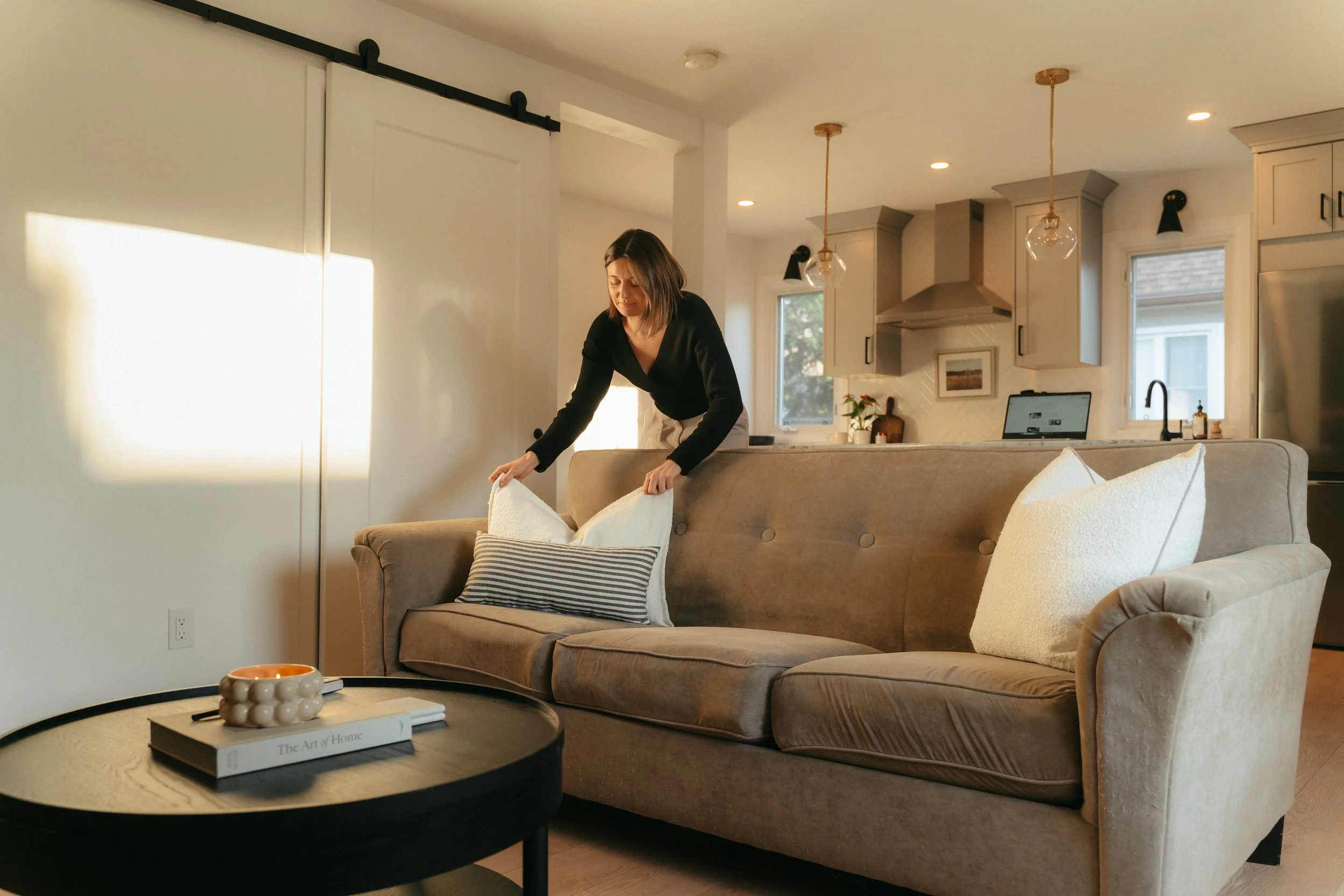 A woman making a bed in a modern, cozy living room with a sofa, decorative pillows, a coffee table, and a kitchen in the background.
