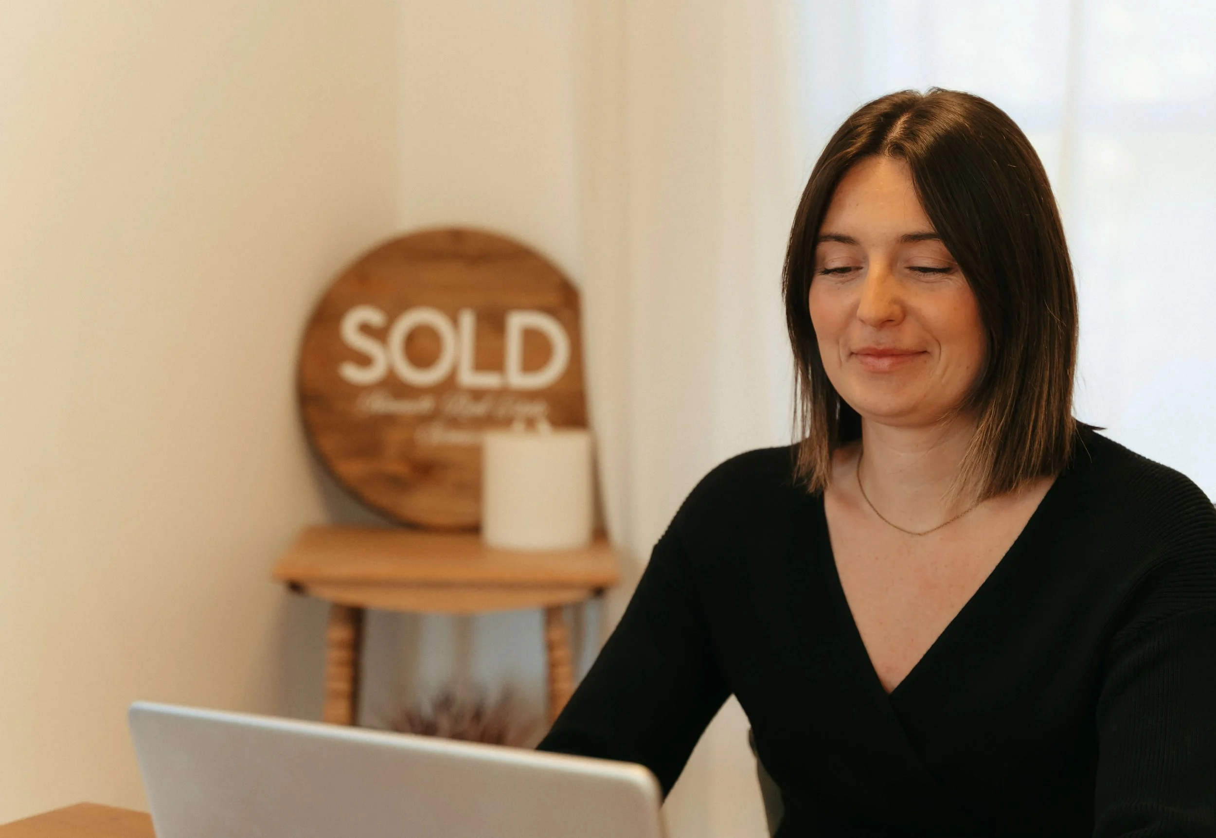 A woman with shoulder-length brown hair and a black top, sitting in front of a laptop, with a 'SOLD' sign on a wooden stand in the background.
