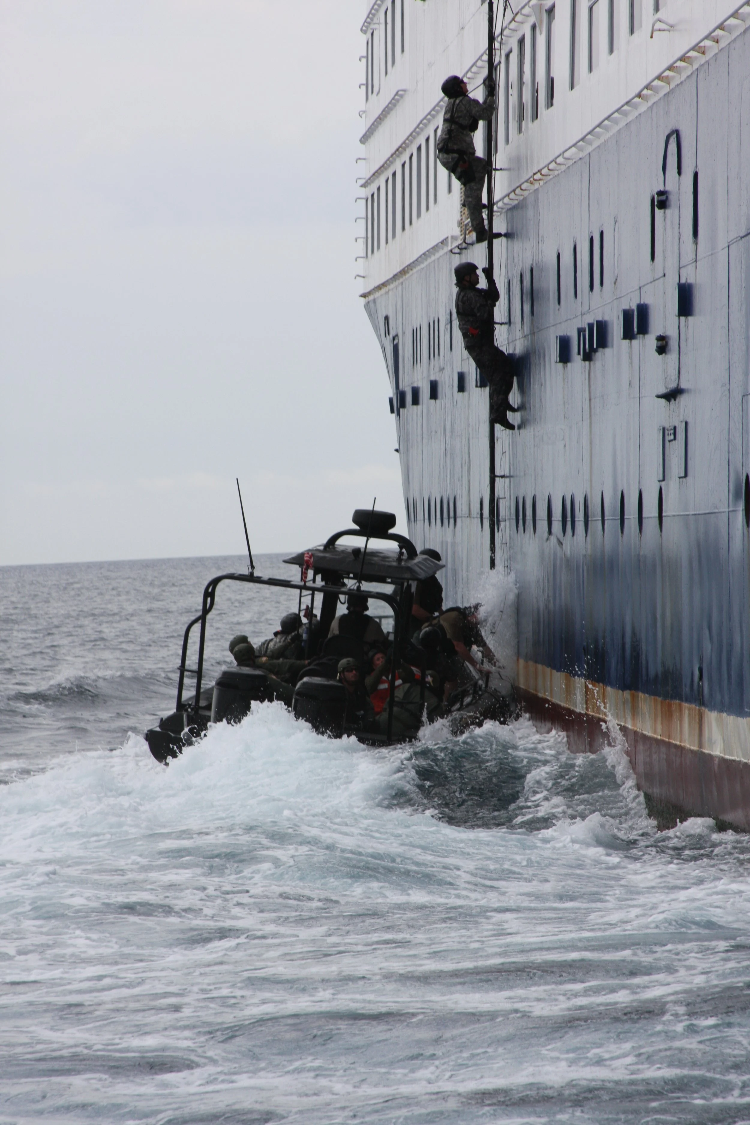 Military personnel and crew members on a boat conducting a boarding exercise on a large ship, with soldiers climbing the ship's side using ropes in rough waters.