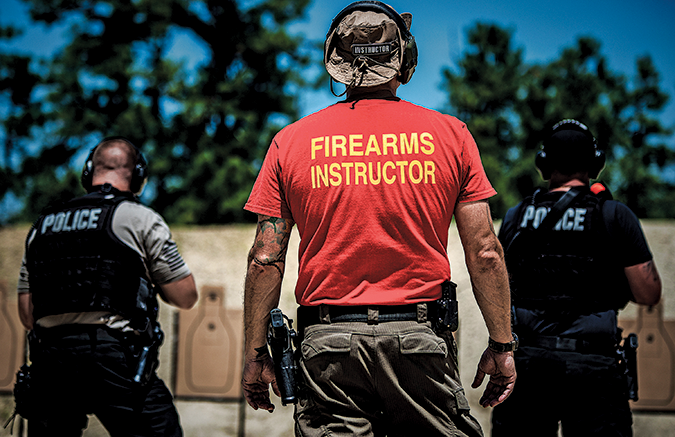 Firearms instructor wearing a red shirt with yellow text, accompanied by police officers at a shooting range with targets in the background.