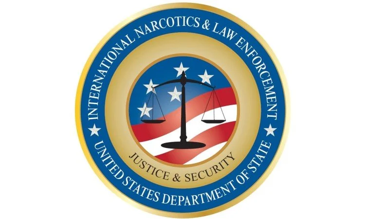 Seal of the United States Department of State for the International Narcotics & Law Enforcement, Justice & Security division, featuring a balanced scale against an American flag background.