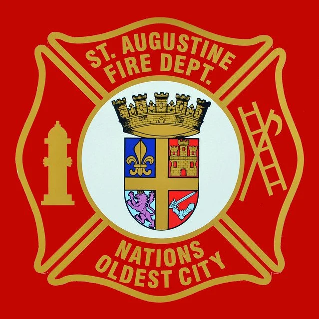 Emblem of the St. Augustine Fire Department with a red background, featuring a shield with a crown, a fleur-de-lis, a castle, a lion, and a firefighter's hook, and text reading 'St. Augustine Fire Dept.', 'Nations Oldest City'.