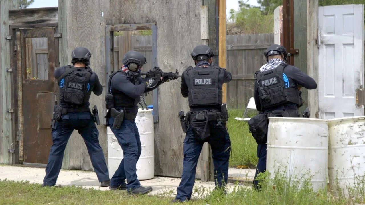 Four police officers in tactical gear conducting a training exercise with one officer aiming a rifle through a door opening in a wooden wall.
