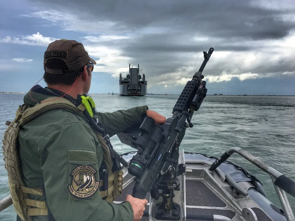 A law enforcement officer in tactical gear on a boat, aiming a mounted firearm at a large ship in the water under cloudy skies.