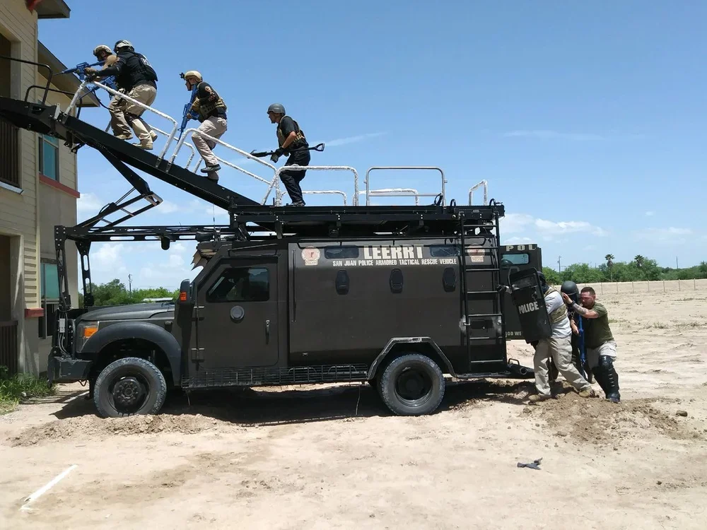 Police officers and rescue personnel are using an armored tactical rescue vehicle to perform a rescue operation, with some officers helping an individual out of the vehicle while others ascend a ladder.