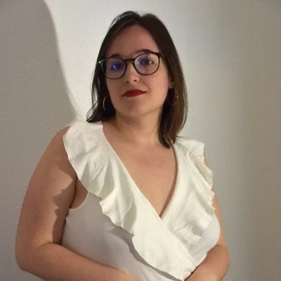 Jeune femme avec lunettes, cheveux bruns, portant un haut blanc avec des volants, se tient contre un mur blanc.
