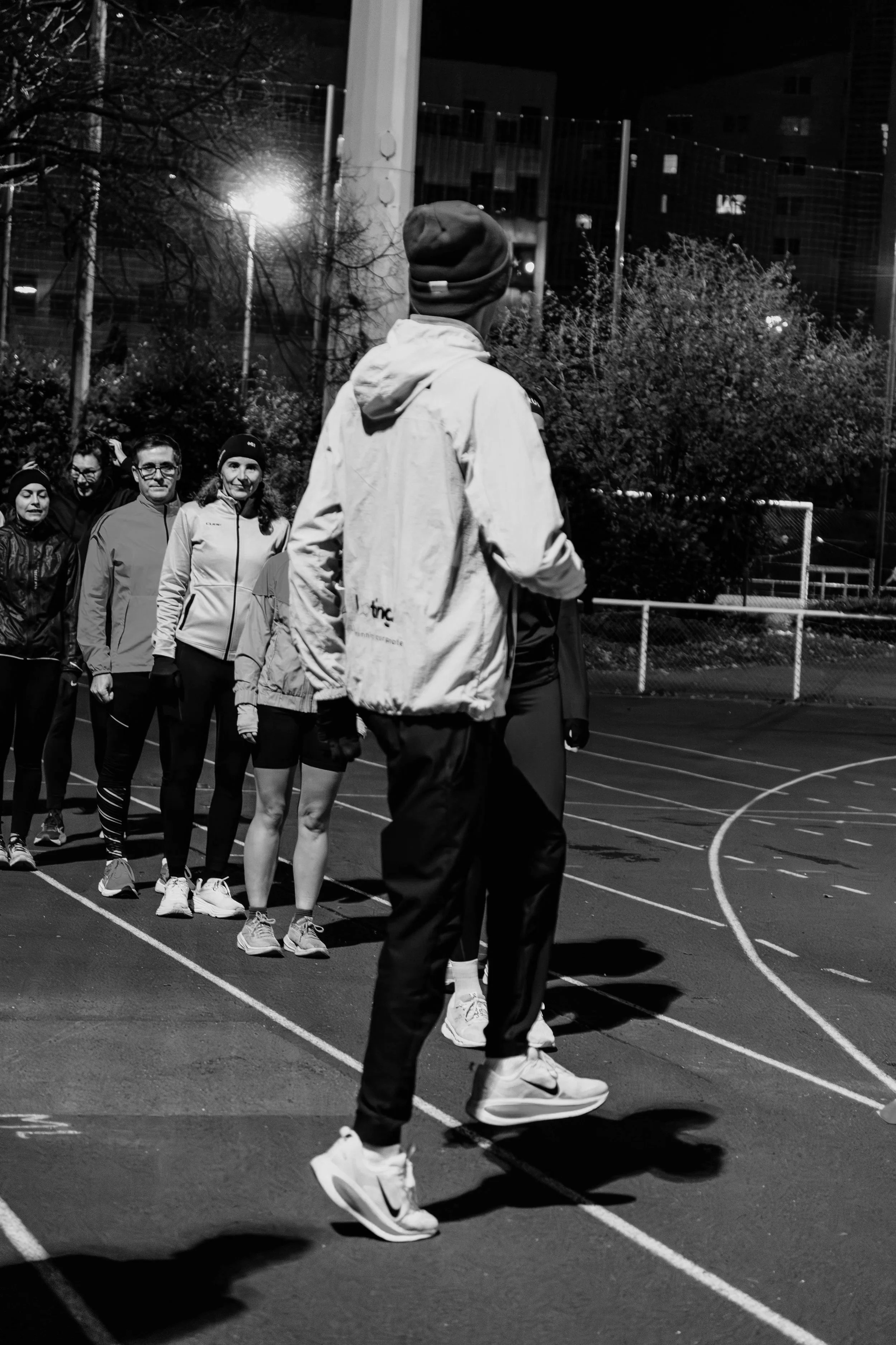 Groupe de personnes attendant dans une file à une séance d'entraînement ou une activité sportive sur une piste en soirée, en plein air, avec des bâtiments et des arbres en arrière-plan.