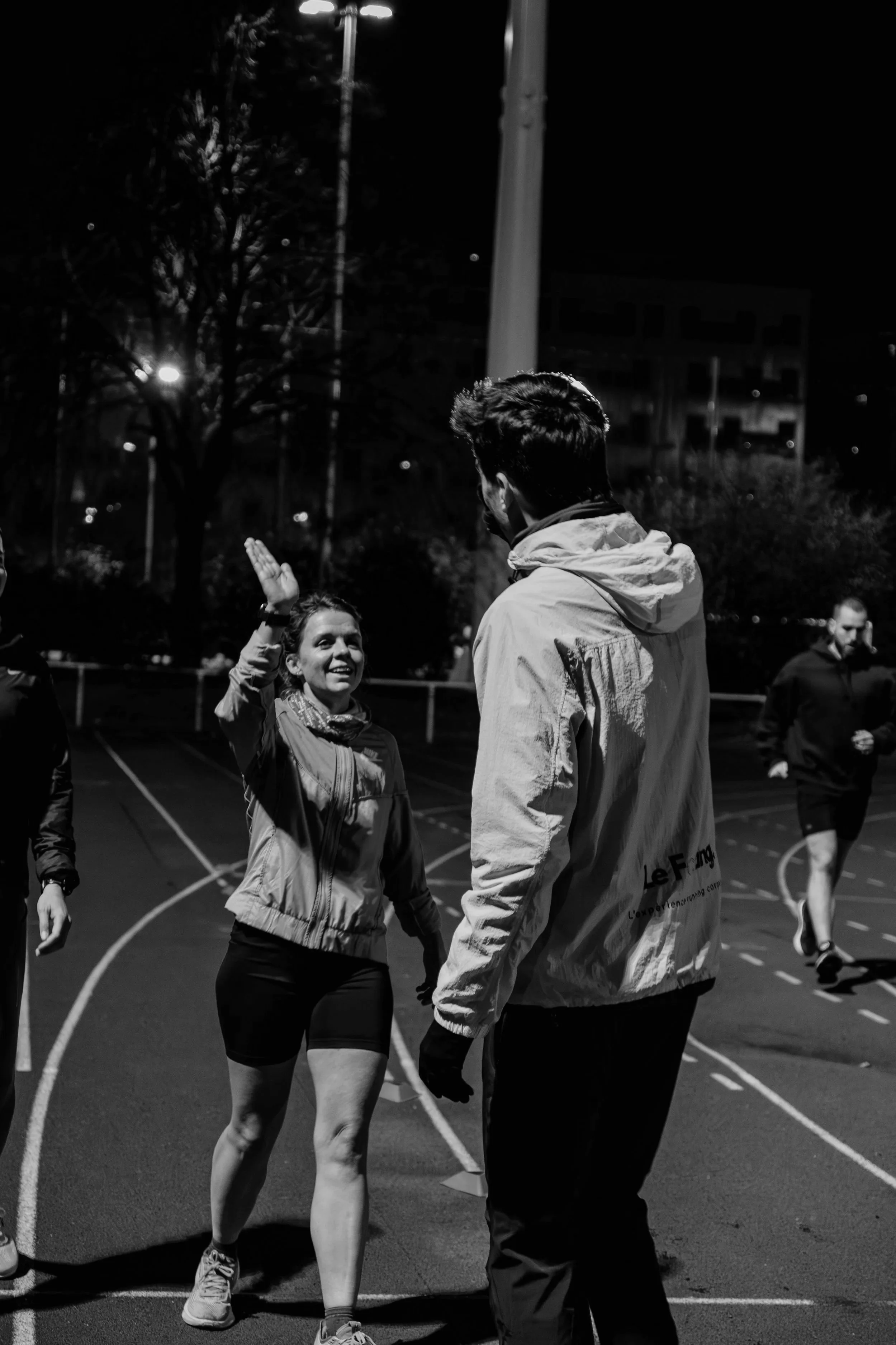 Deux personnes discutent sur une piste d'athlétisme la nuit, un homme et une femme, la femme sourit et semble saluer l'homme.
