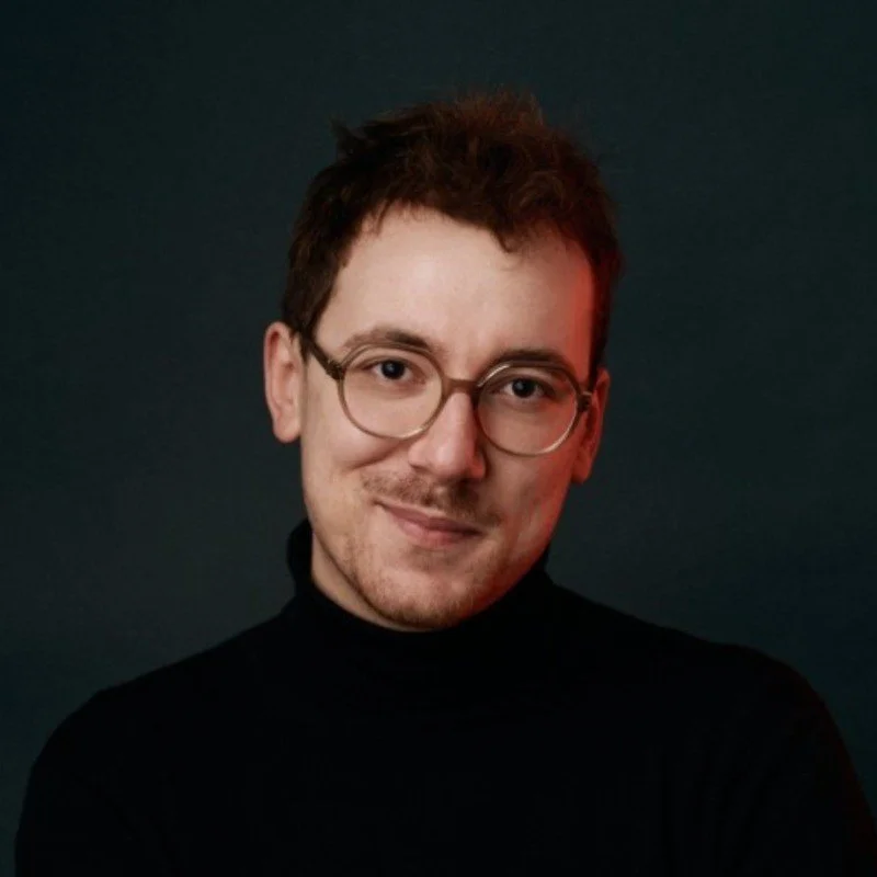 Portrait d'un jeune homme avec lunettes, souriant, portant un pull noir, fond sombre.