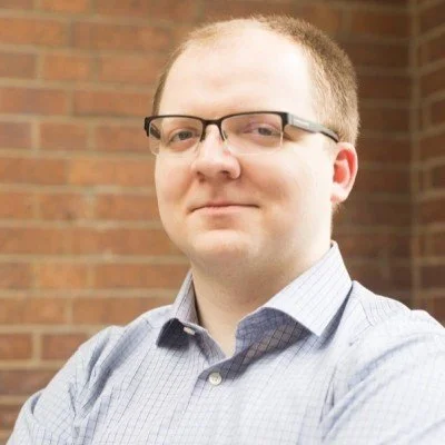 A man with glasses and short hair wearing a collared shirt standing outdoors in front of a brick wall.