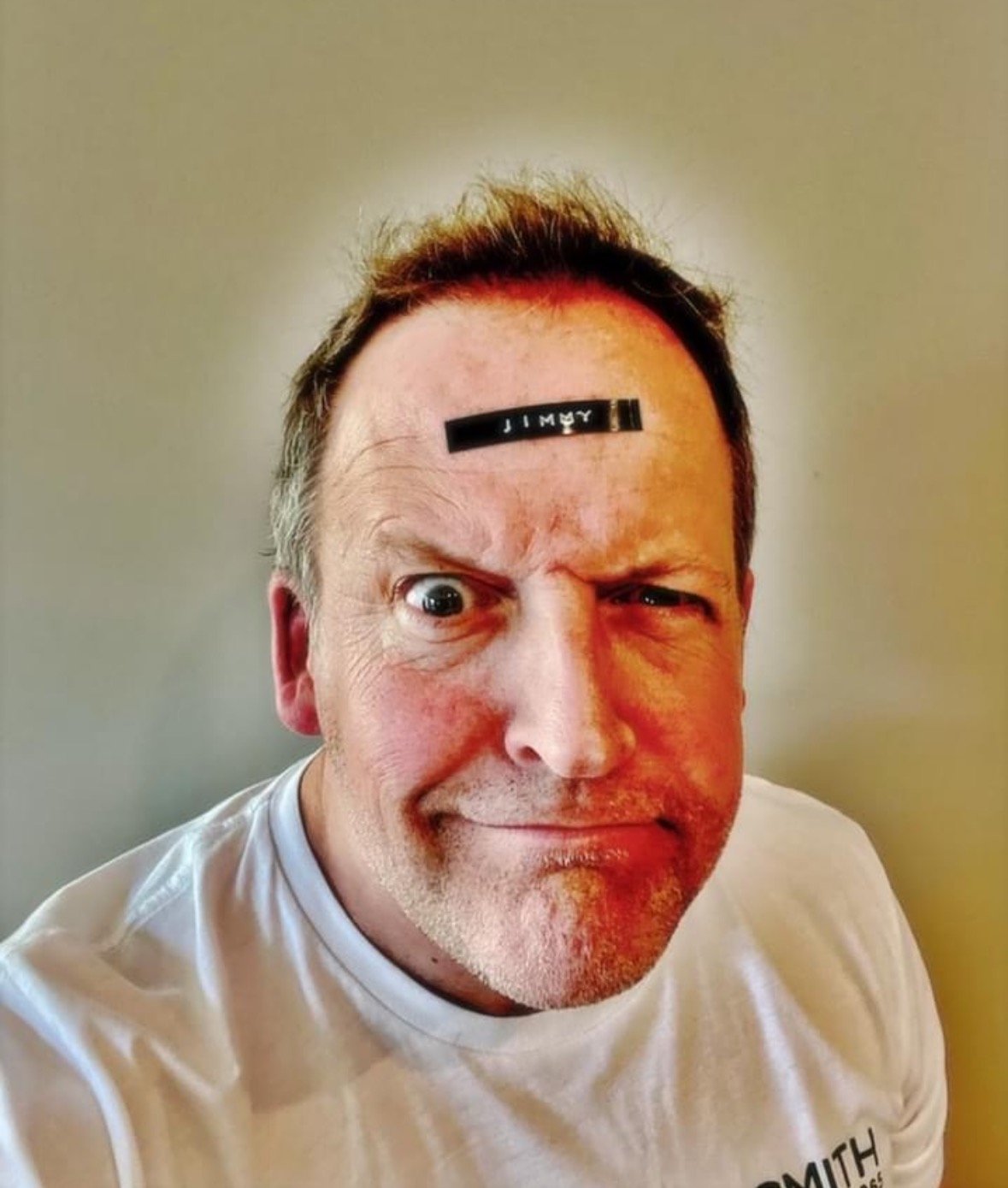 A man with one eye winking, a sticker on his forehead that reads 'JIMMY', wearing a white T-shirt, smiling with a playful expression.