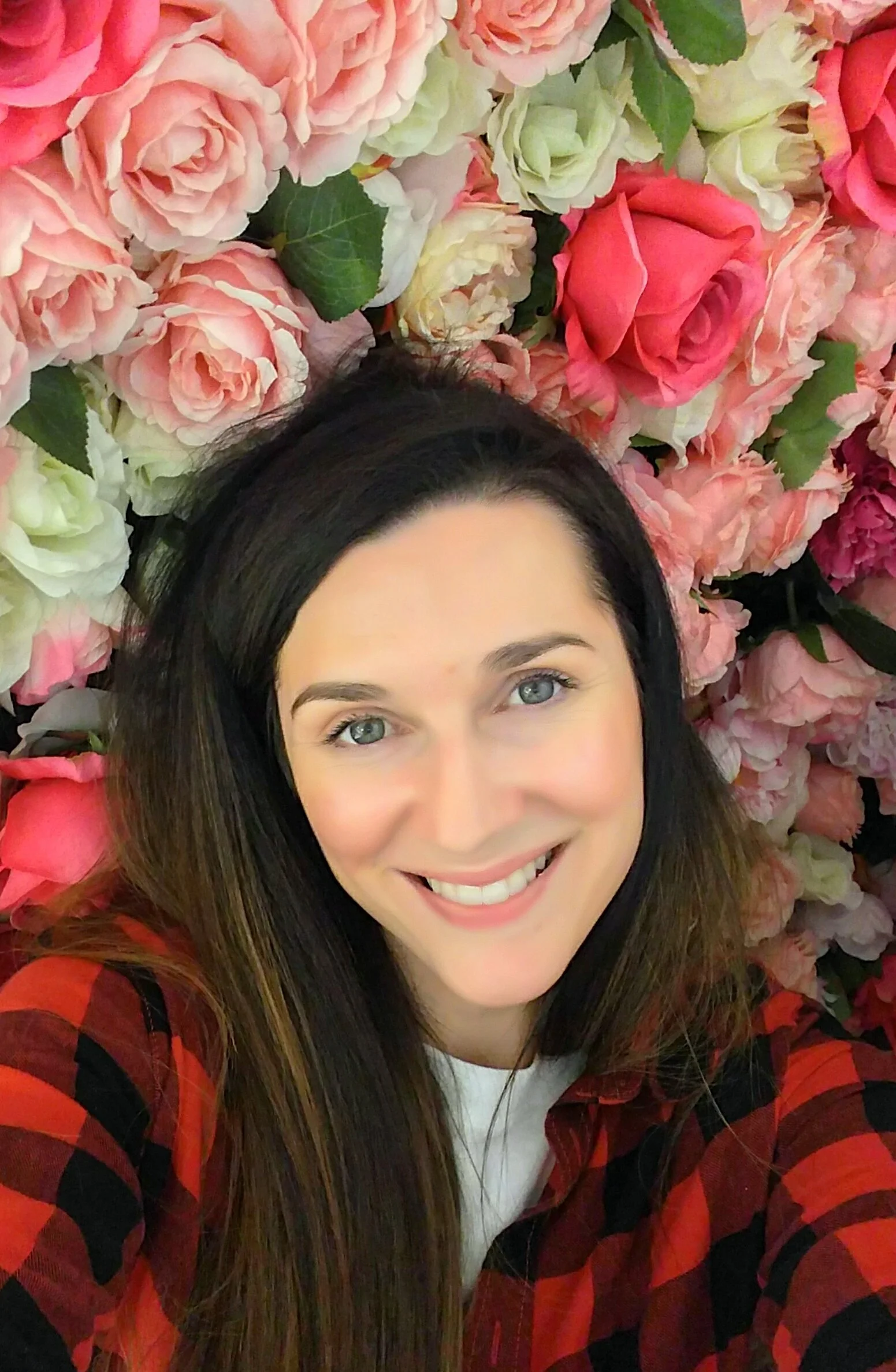 A woman with long dark hair, blue eyes, and a bright smile taking a selfie in front of a backdrop of pink and white flowers, wearing a red and black plaid shirt.