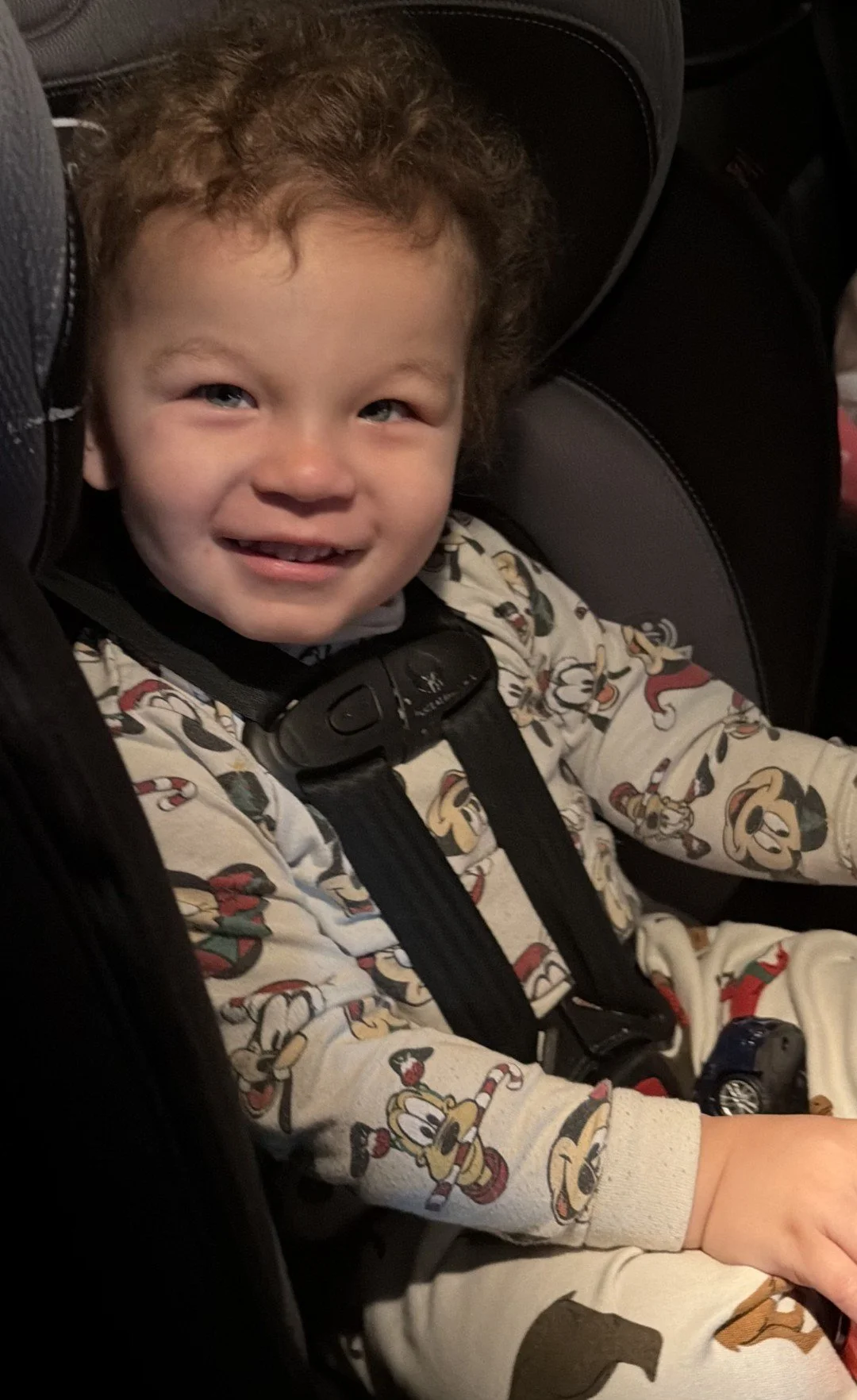 A young child with curly hair smiling while seated in a car seat, wearing a Mickey Mouse themed outfit.