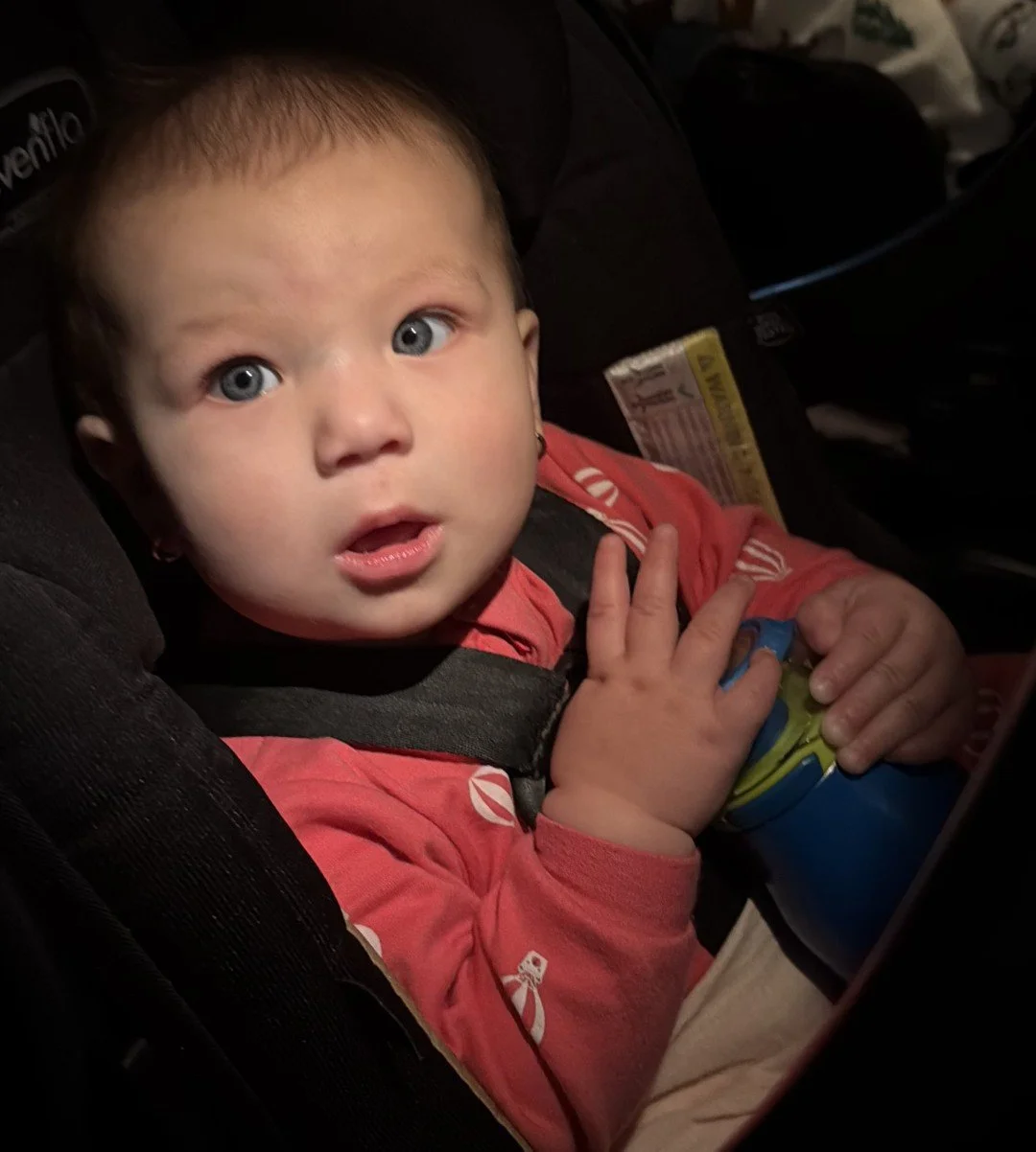A young child with blue eyes and light brown hair sitting in a car seat with a surprised or curious expression, holding a blue and green sippy cup.