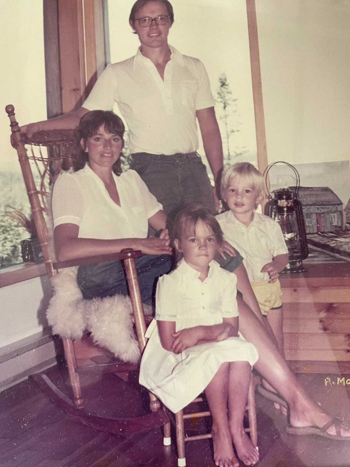 A family portrait with two adults and three children inside a room, with a window showing trees in the background. The woman is sitting on a wooden rocking chair, and the man is standing behind her. The children are sitting on or near the woman's lap. The scene has vintage, slightly faded colors.
