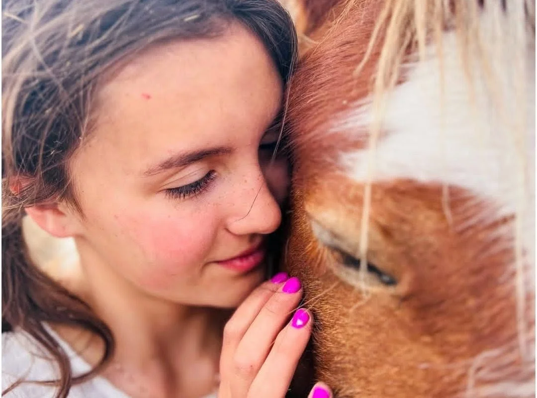 A young woman with closed eyes and pink nails gently touching a horse's nose.