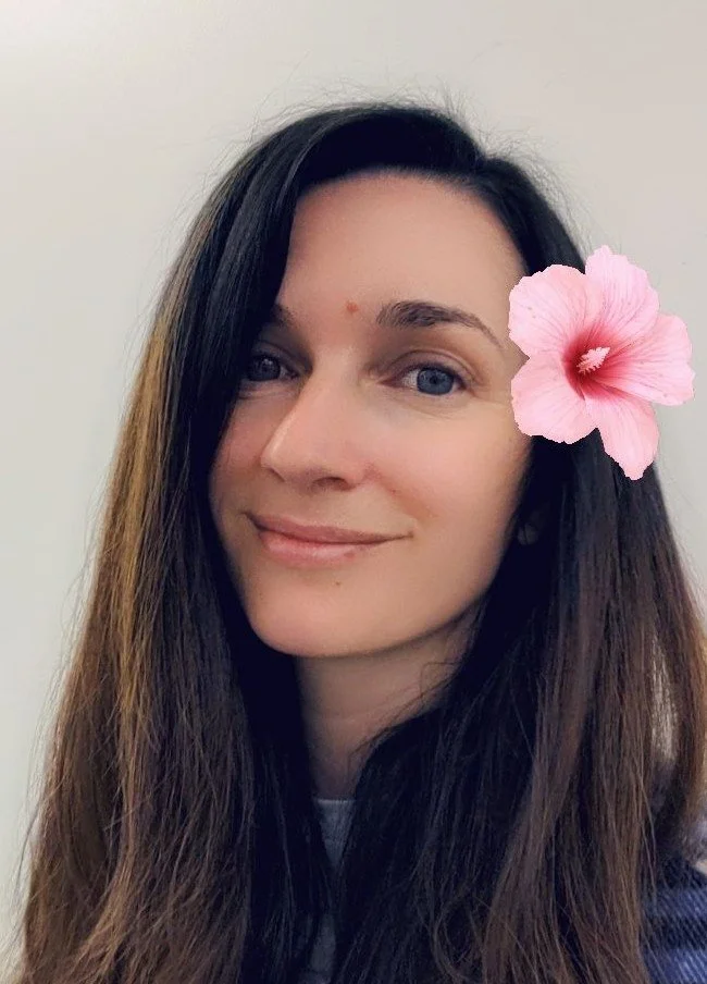 A woman with long dark hair and blue eyes smiling, with a pink hibiscus flower in her hair.