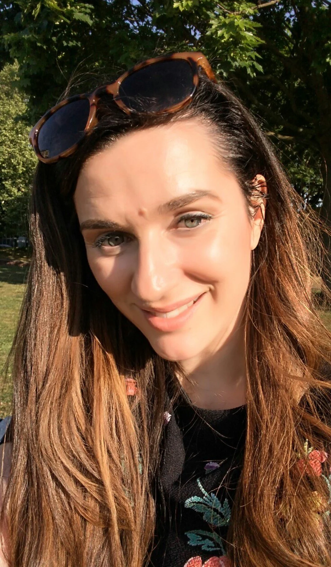 A woman with long brown hair, sunglasses on her head, smiling outdoors in sunlight with trees in the background.