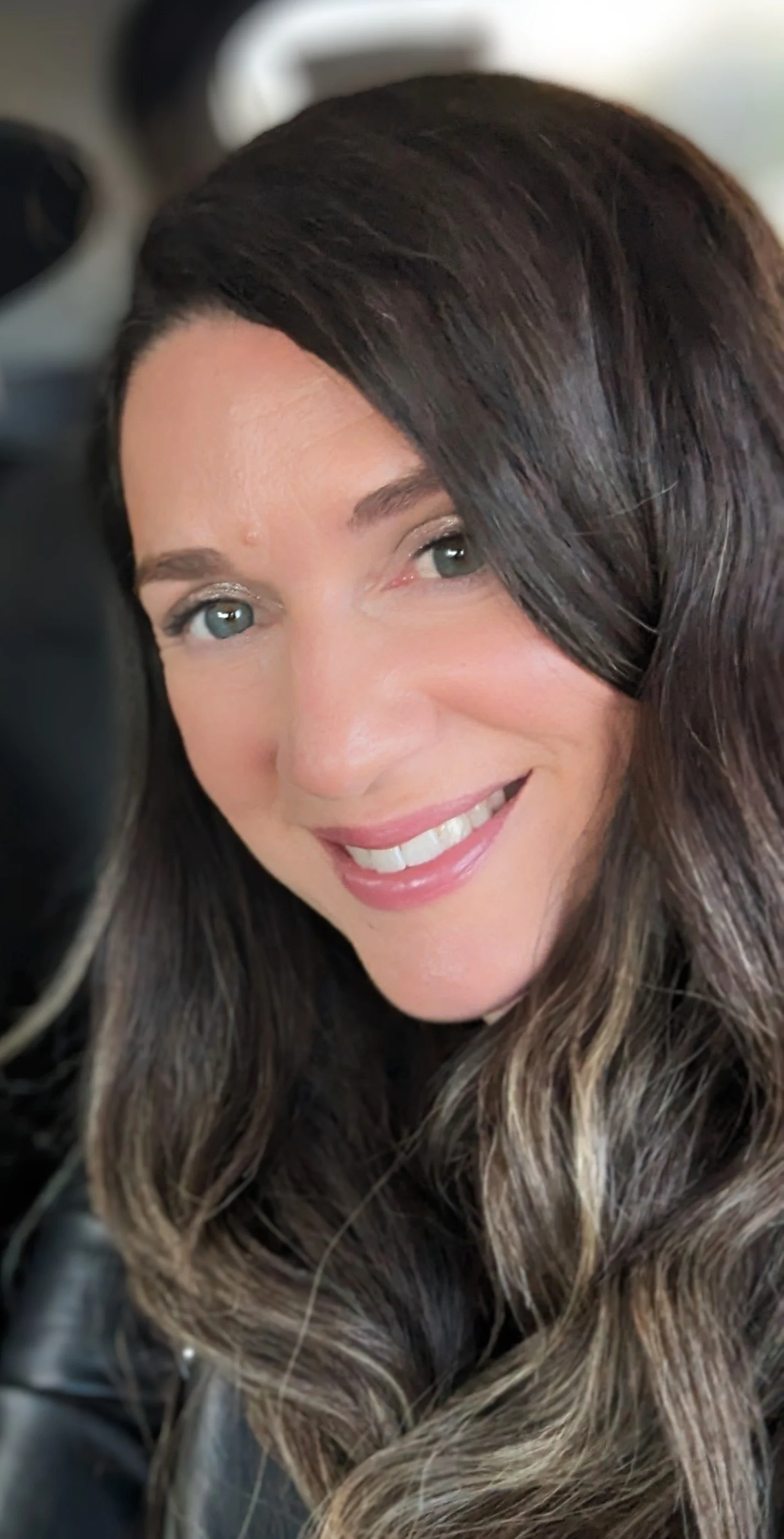 Close-up of a smiling woman with wavy brown hair and fair skin, inside a vehicle.