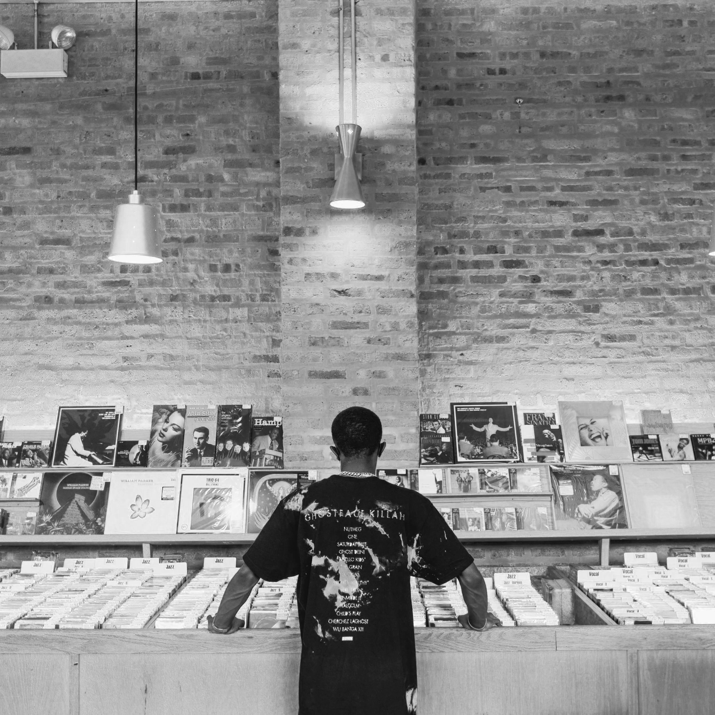 A person browsing vinyl records in a record store with a brick wall background and modern hanging lights.