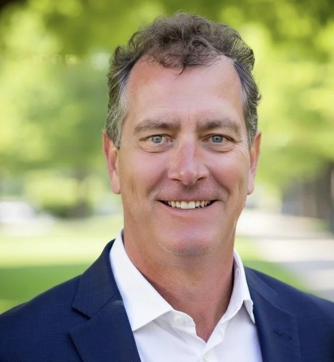 Headshot of a man with gray hair and blue eyes wearing a navy blazer and white shirt, standing outdoors with greenery in the background.