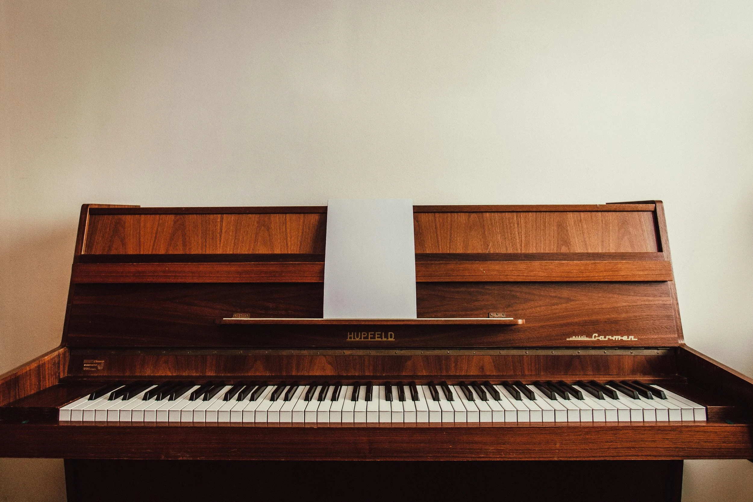 A vintage wooden upright piano with a blank sheet of music on the music stand against a plain wall.