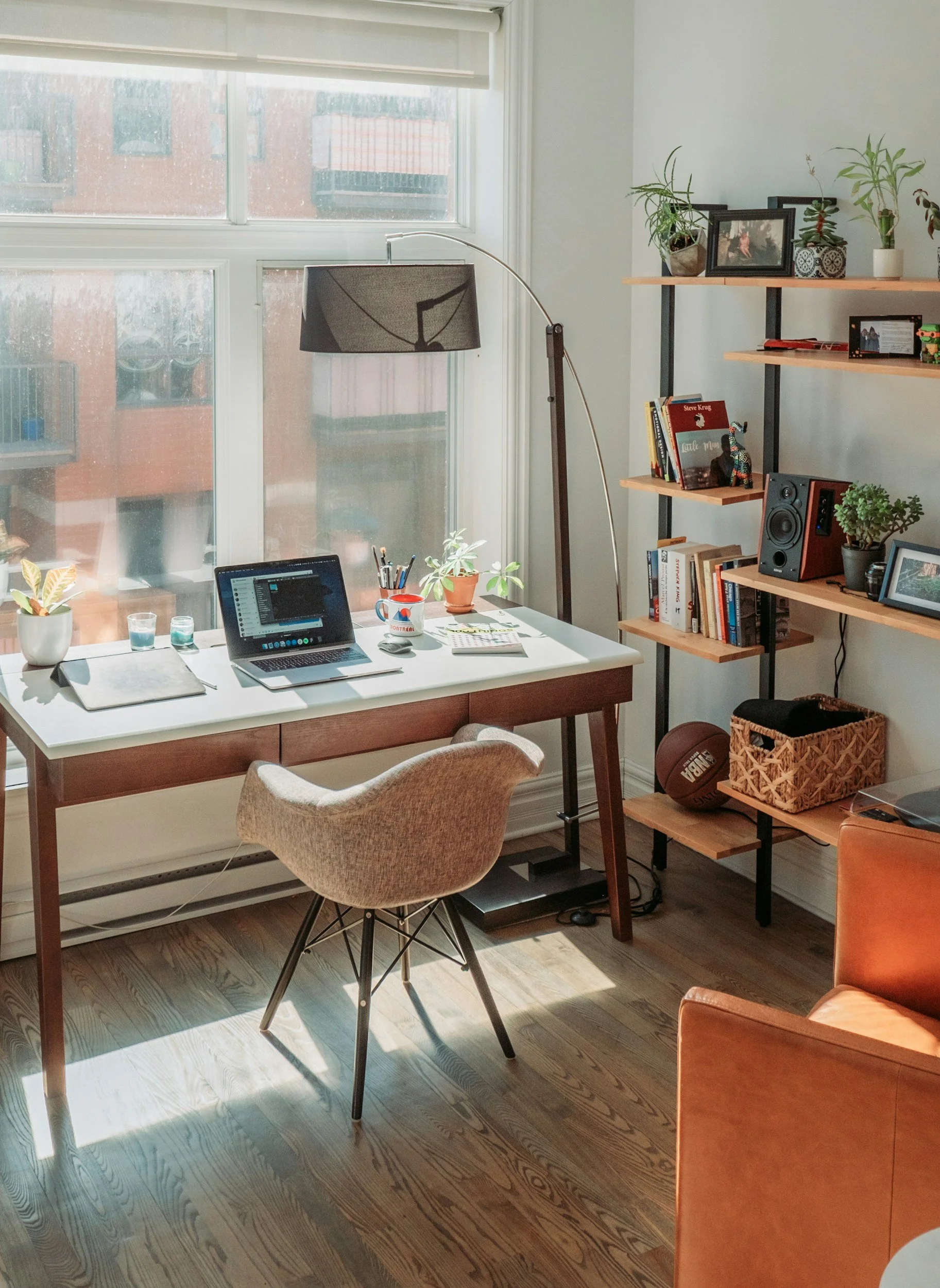 A cozy home office with a white desk near a large window, a laptop, potted plants, and a beige chair. Bookshelves contain books, framed photos, and plants. A basketball and a woven basket sit on the floor.