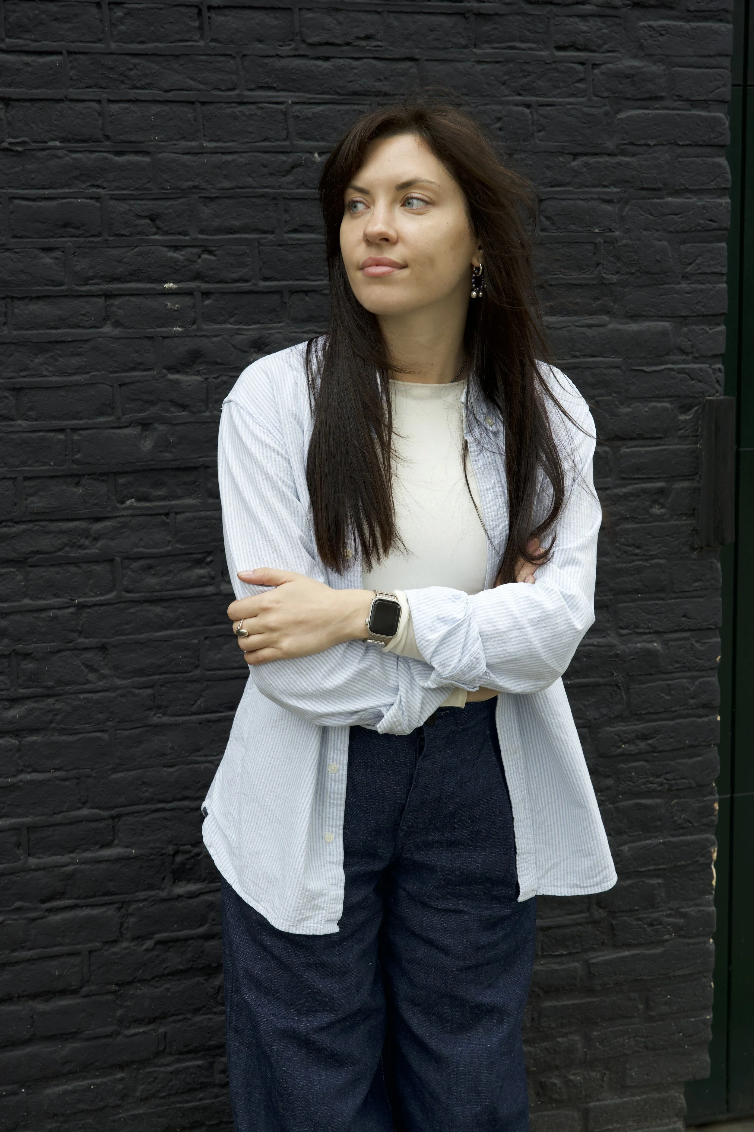 Woman with long dark hair, wearing a white shirt, light blue striped jacket, dark jeans, and a smartwatch, standing against a black brick wall.