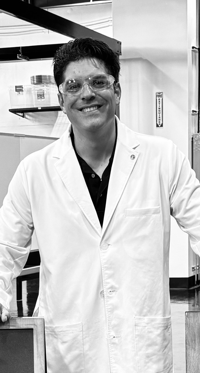A man wearing glasses and a white lab coat, smiling in a laboratory setting.