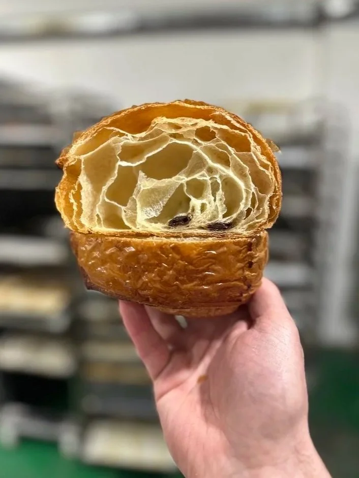 A hand holding a large, partially cut open pain au chocolat with an airy, flaky interior.