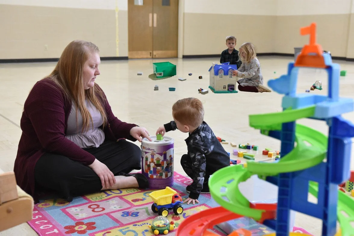 Children playing with toys on a colorful carpet in an indoor play area, with a woman sitting nearby and a large plastic toy structure in the foreground.