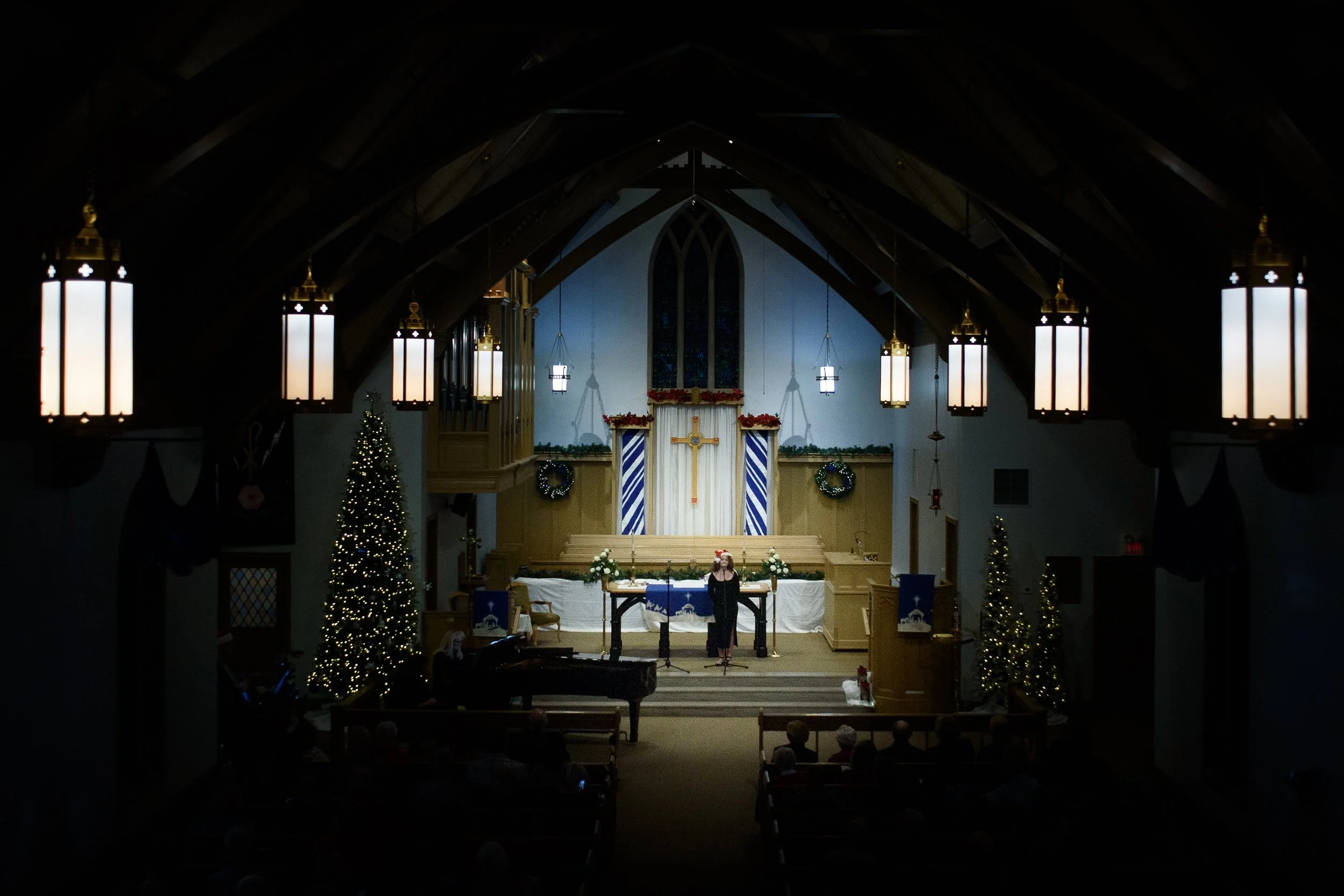 Interior of a church decorated for Christmas with Christmas trees and wreaths, featuring a woman standing at a microphone stage with a piano nearby, illuminated by hanging lantern lights.