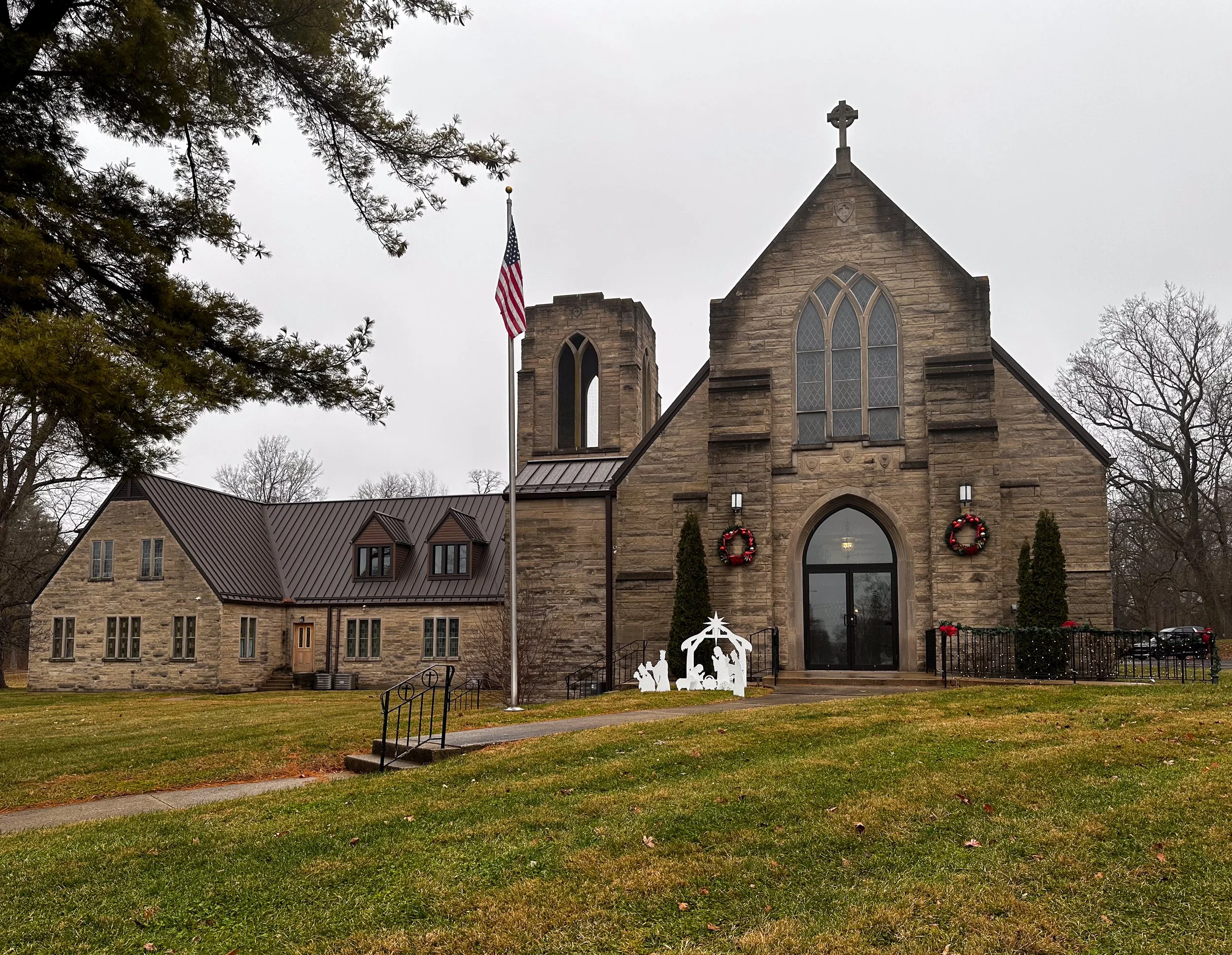 A stone church decorated with Christmas wreaths and holiday decorations on the front porch, with a nativity scene in front. The church has large gothic-style windows, an American flag on a flagpole, and a tall bell tower. The surrounding yard is grassy with trees around, and the sky is overcast.