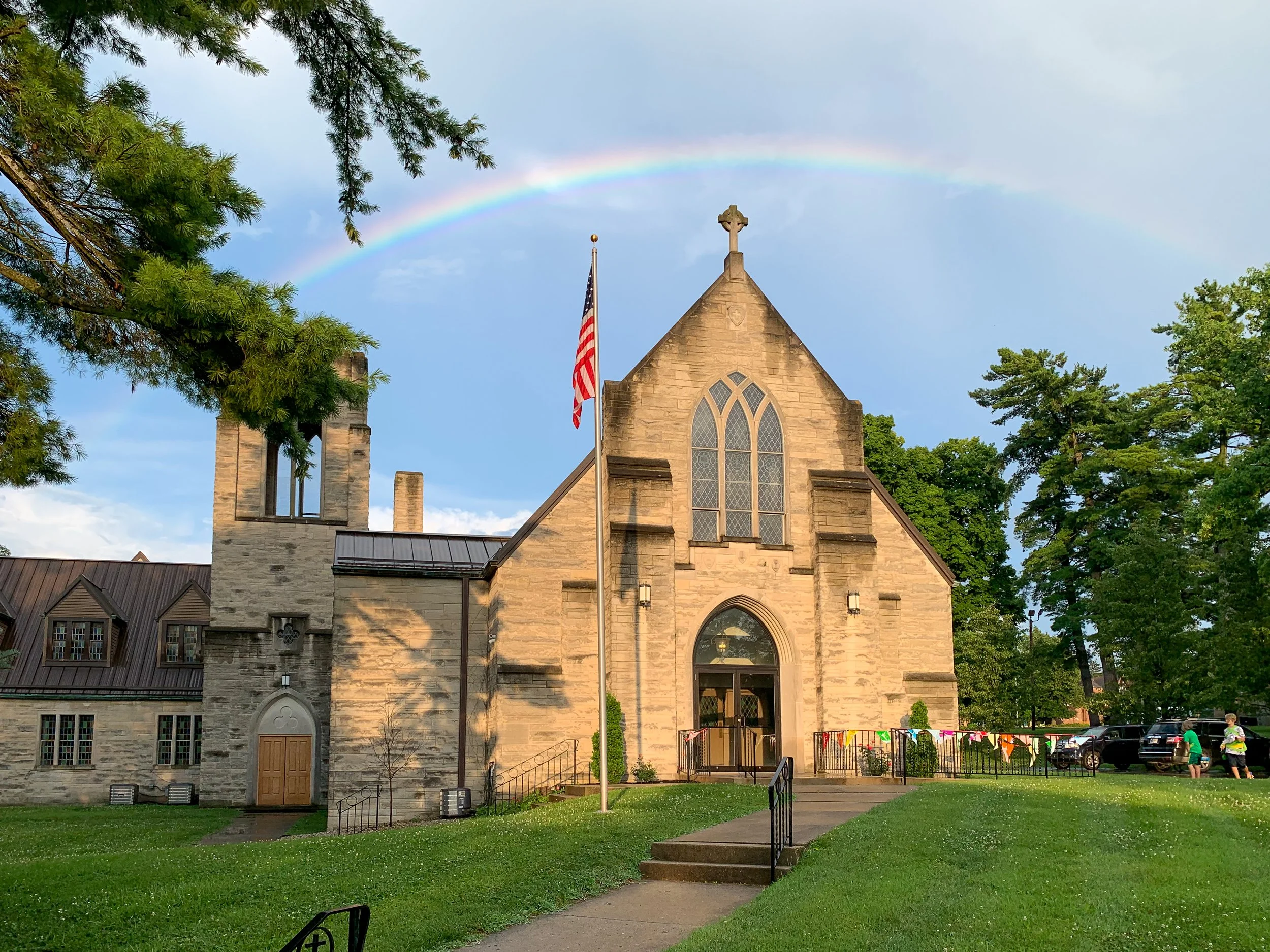 A stone church with large arched windows and a cross on top, an American flag in front, and a rainbow in the sky above, surrounded by green trees and a well-maintained lawn.