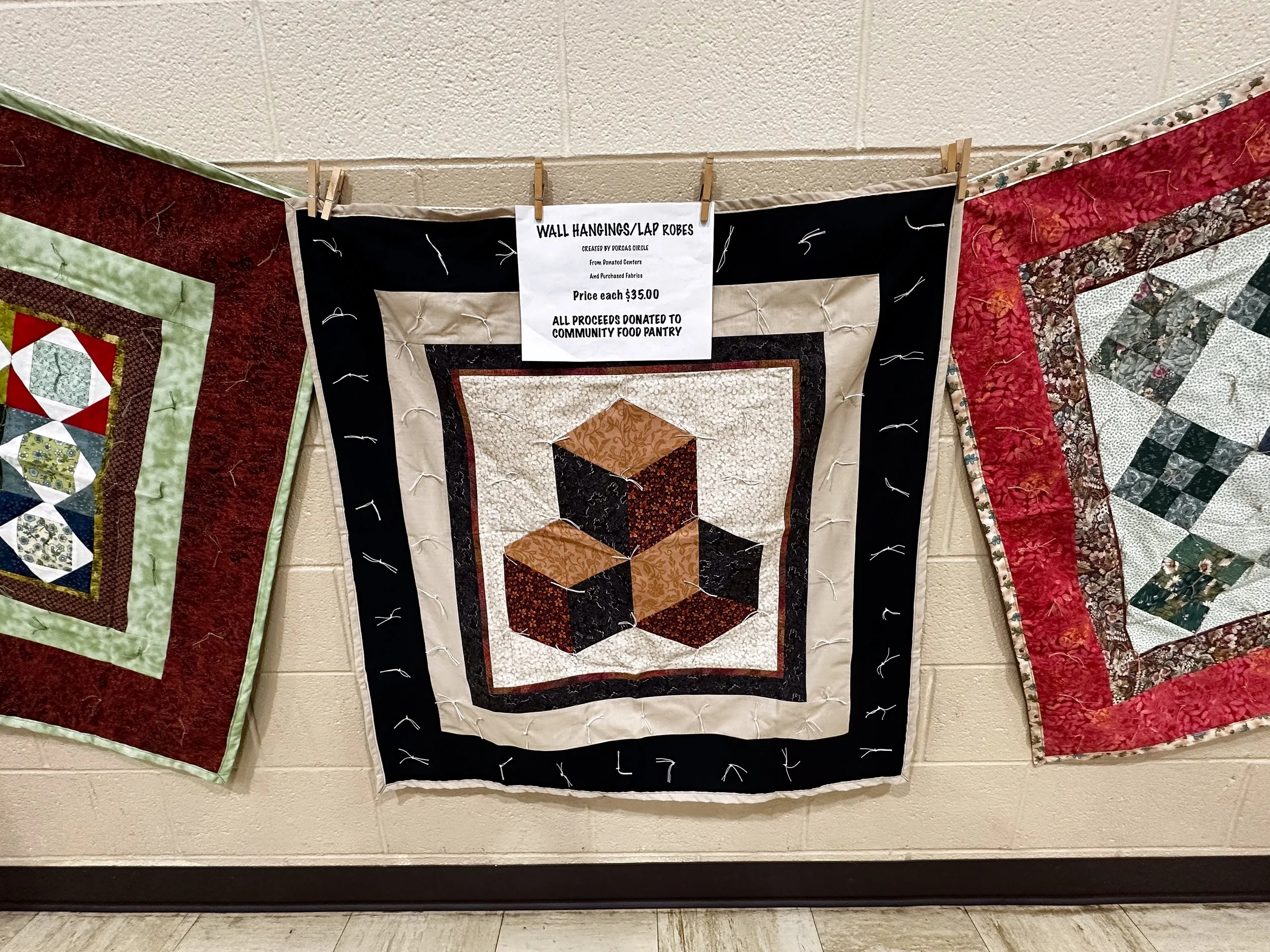 Colorful quilt hanging on a wall with a sign promoting wall hangings lap robes for sale, priced at $35 each, with all proceeds benefiting the community food pantry.