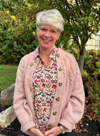An older woman with white hair, glasses, and a warm smile standing outdoors in a garden. She is wearing a pink cardigan over a colorful, patterned blouse.