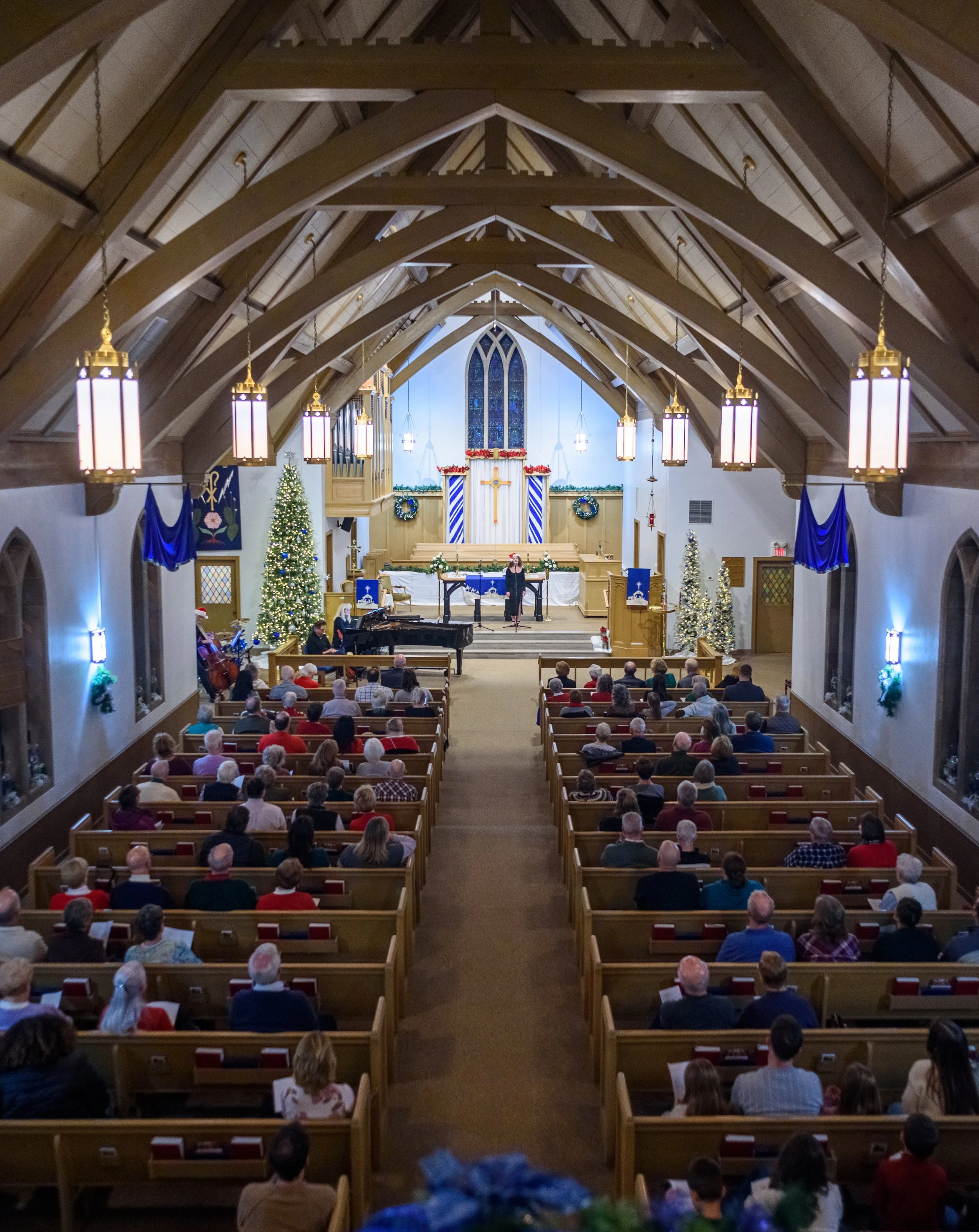 Christmas service in a church with decorated trees, wreaths, and blue curtains; congregation seated in wooden pews facing the altar with a person singing at the microphone.