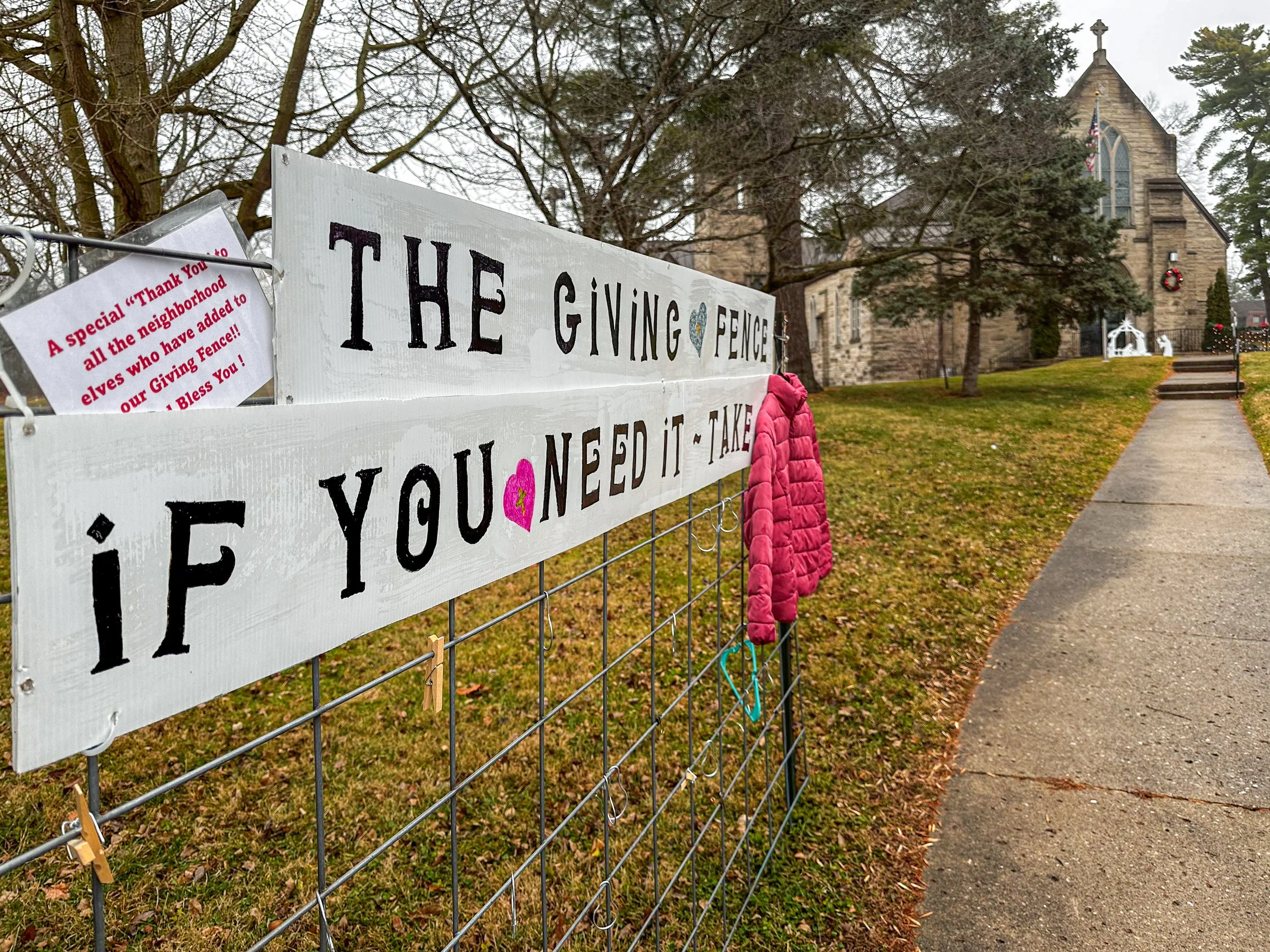 A church courtyard decorated with Christmas wreaths and reindeer figures. In the foreground, a fence holds signs with a Christmas message, including pink scarves hanging on the fence.