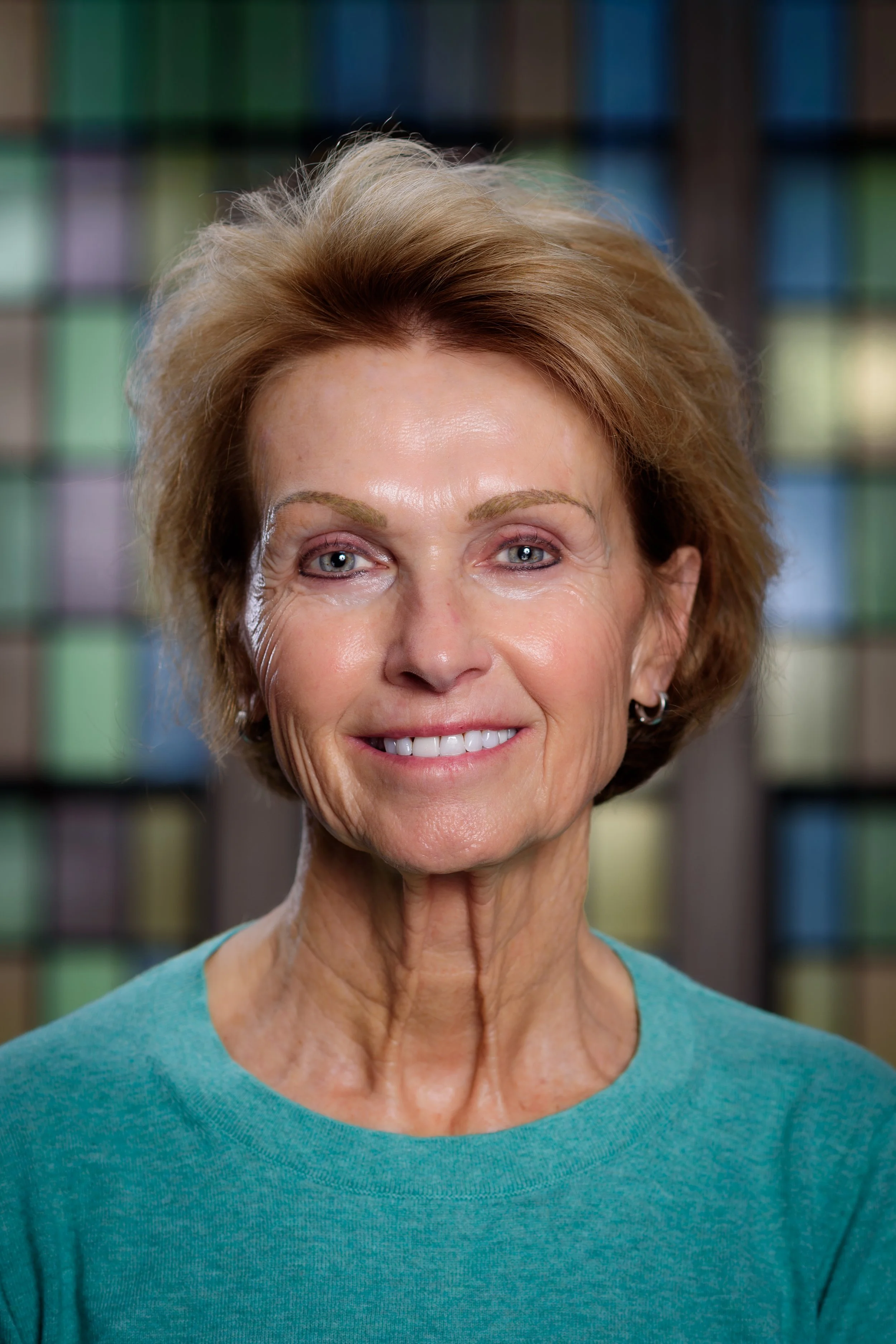 A close-up of a smiling older woman with short, reddish-brown hair, wearing a teal top, standing in front of a colorful, out-of-focus background.