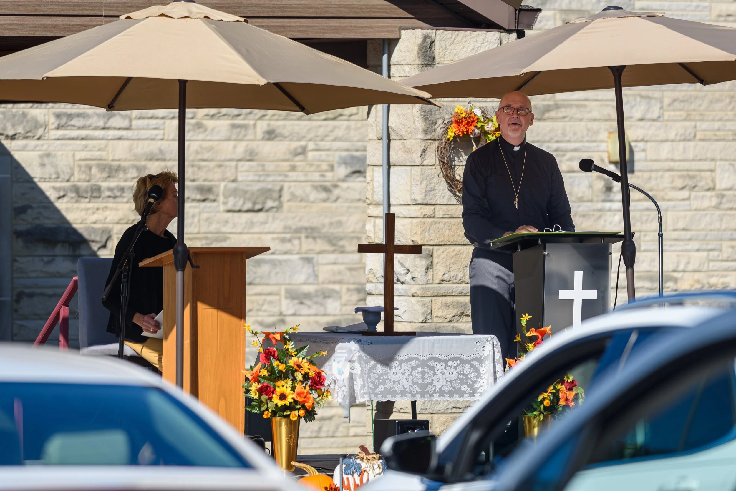A priest and a woman are outdoors at a religious ceremony, standing at podiums with microphones under two large umbrellas. There is a cross and a table with a lace cloth, flowers, and a chalice. The background features a stone wall with a flower wreath.