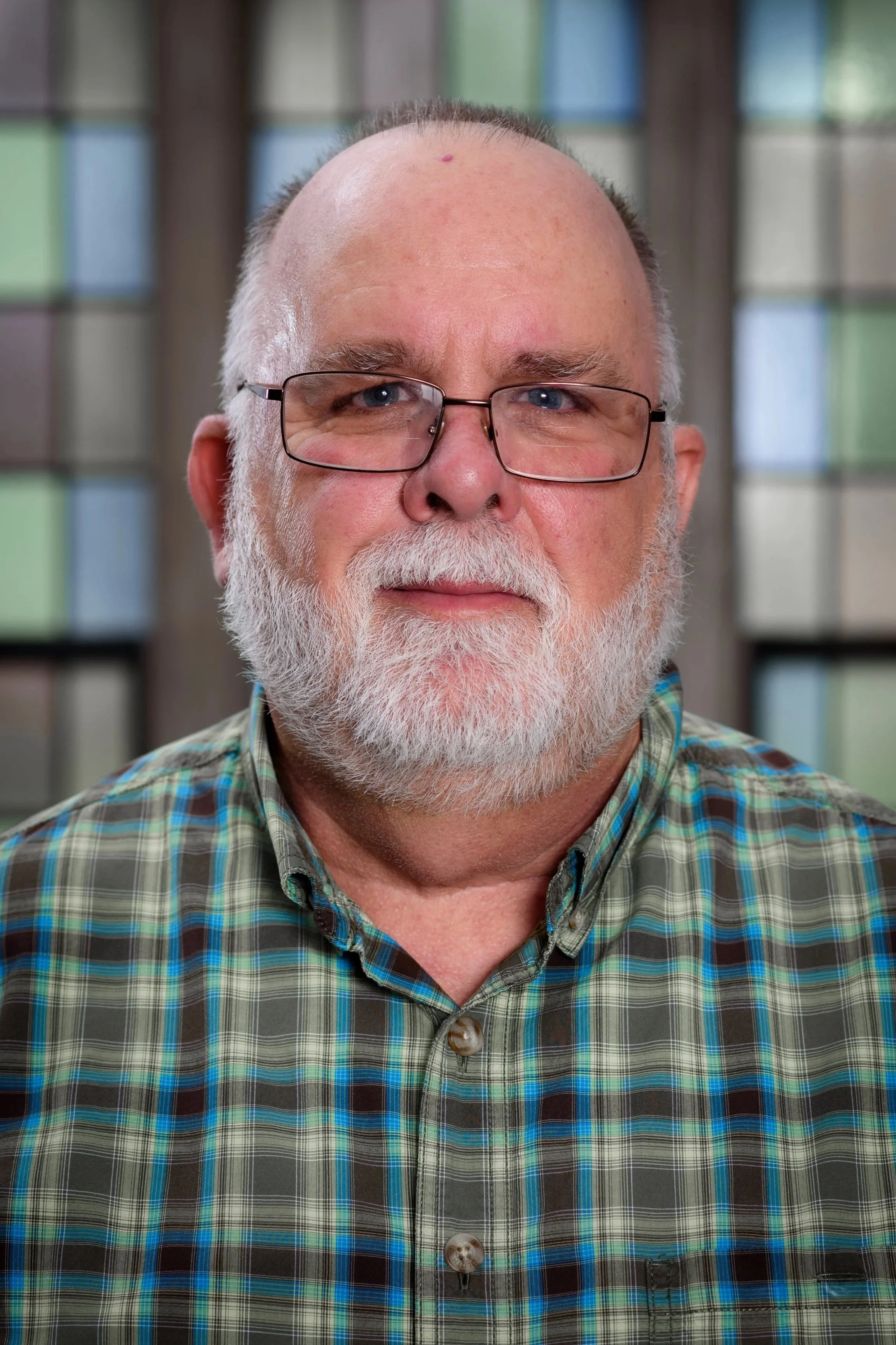 Close-up of an older man with white hair, beard, and glasses wearing a plaid shirt, standing in front of a background with blurred stained glass windows.
