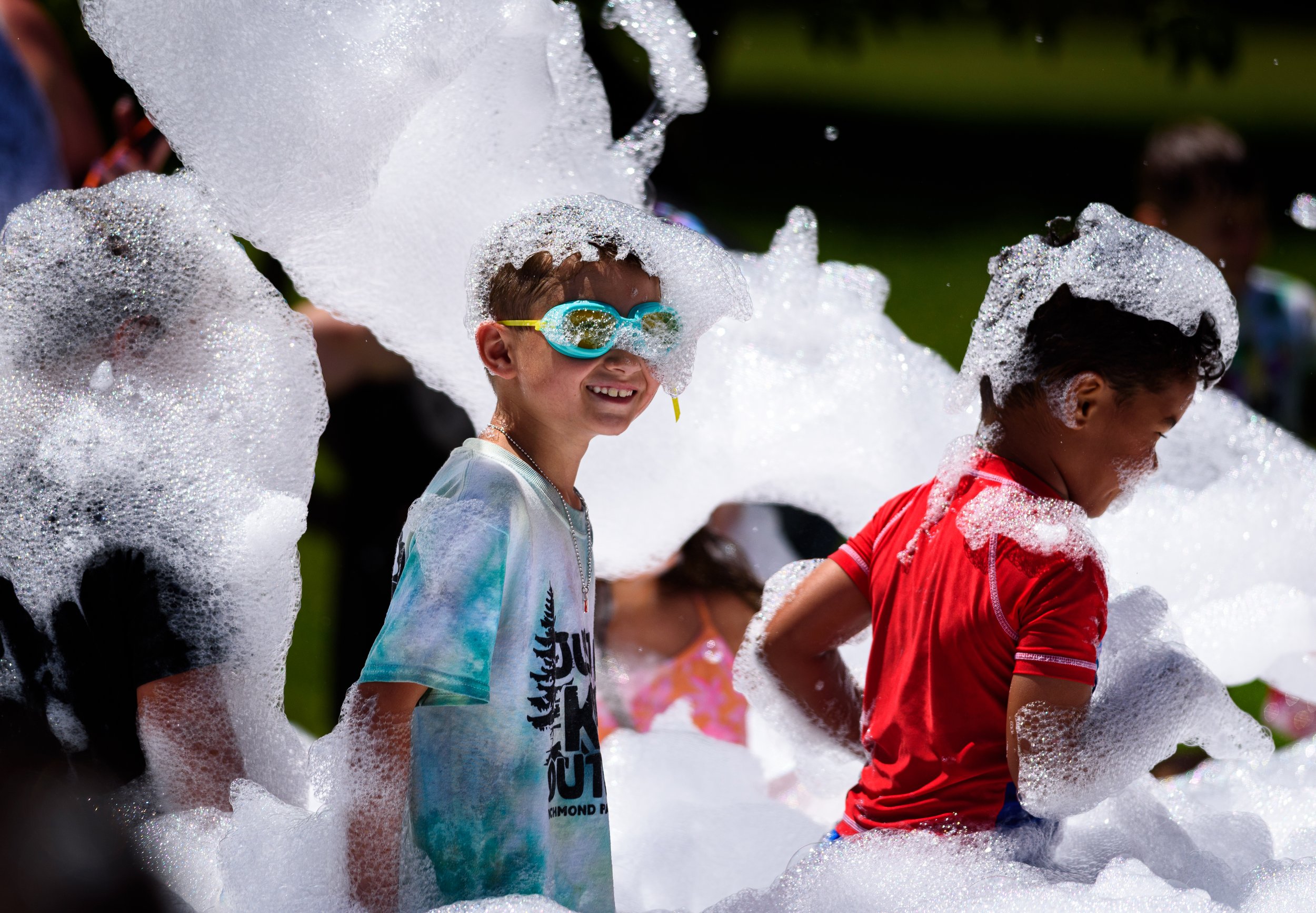 Children playing in a foam party, wearing swimwear and goggles, surrounded by bubbles and soap suds.