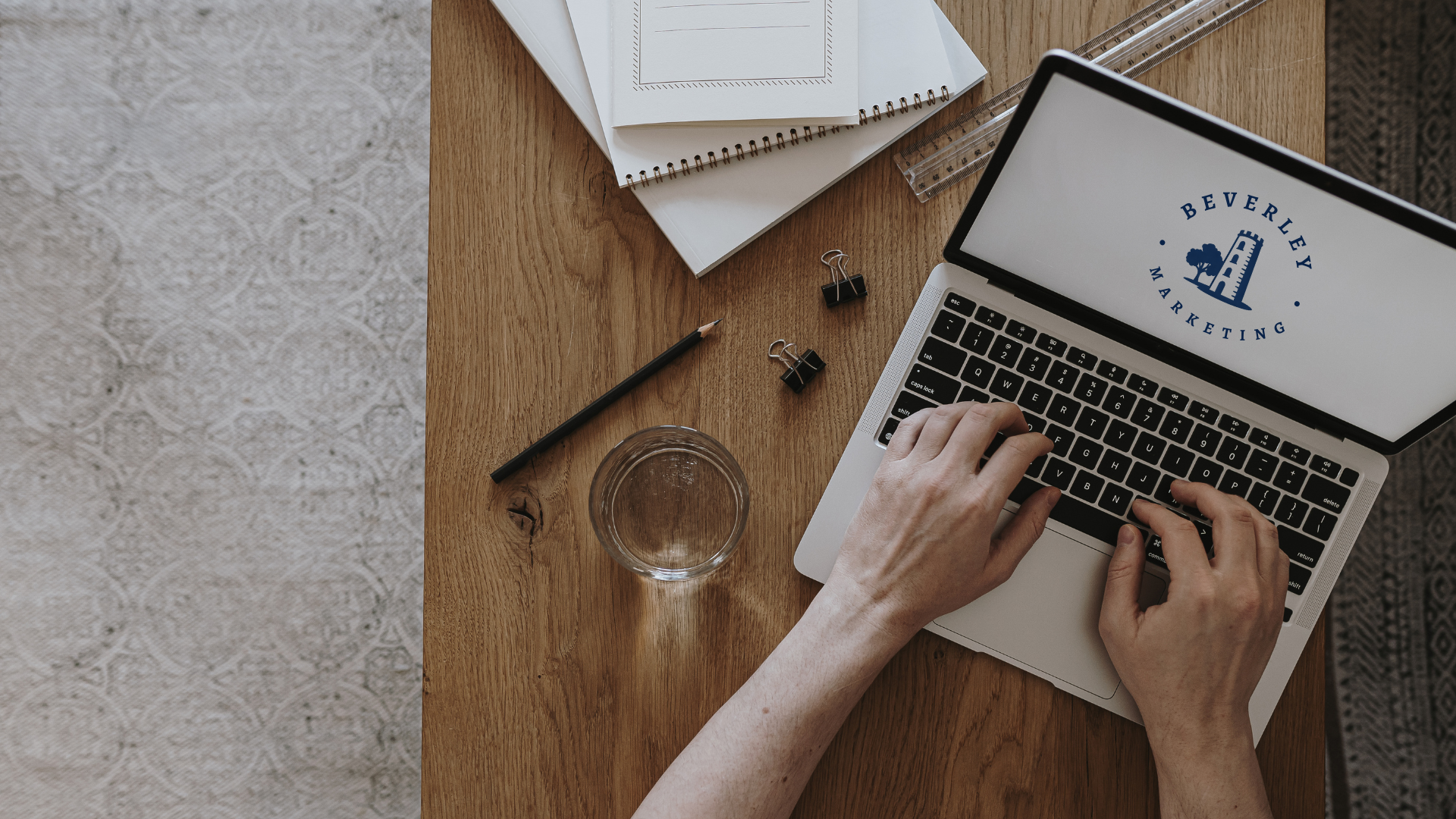 Overhead view of a wooden desk with a person typing on a laptop displaying a Beverley Marketing logo, alongside notebooks, a pen, a glass of water, and office supplies.