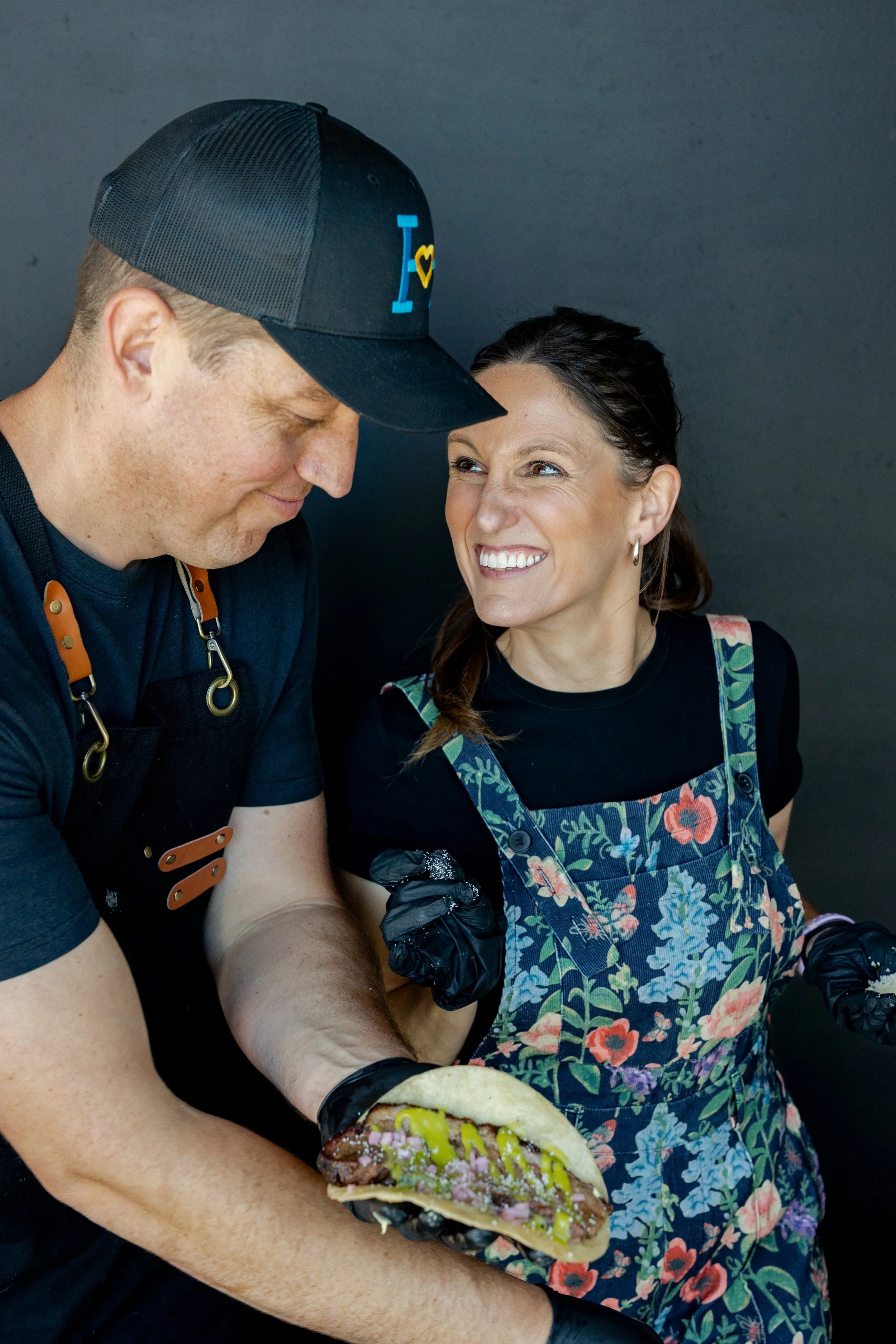 A man and woman sharing a moment as the woman holds a taco filled with meat, vegetables, and sauce, with smiles and close interaction.